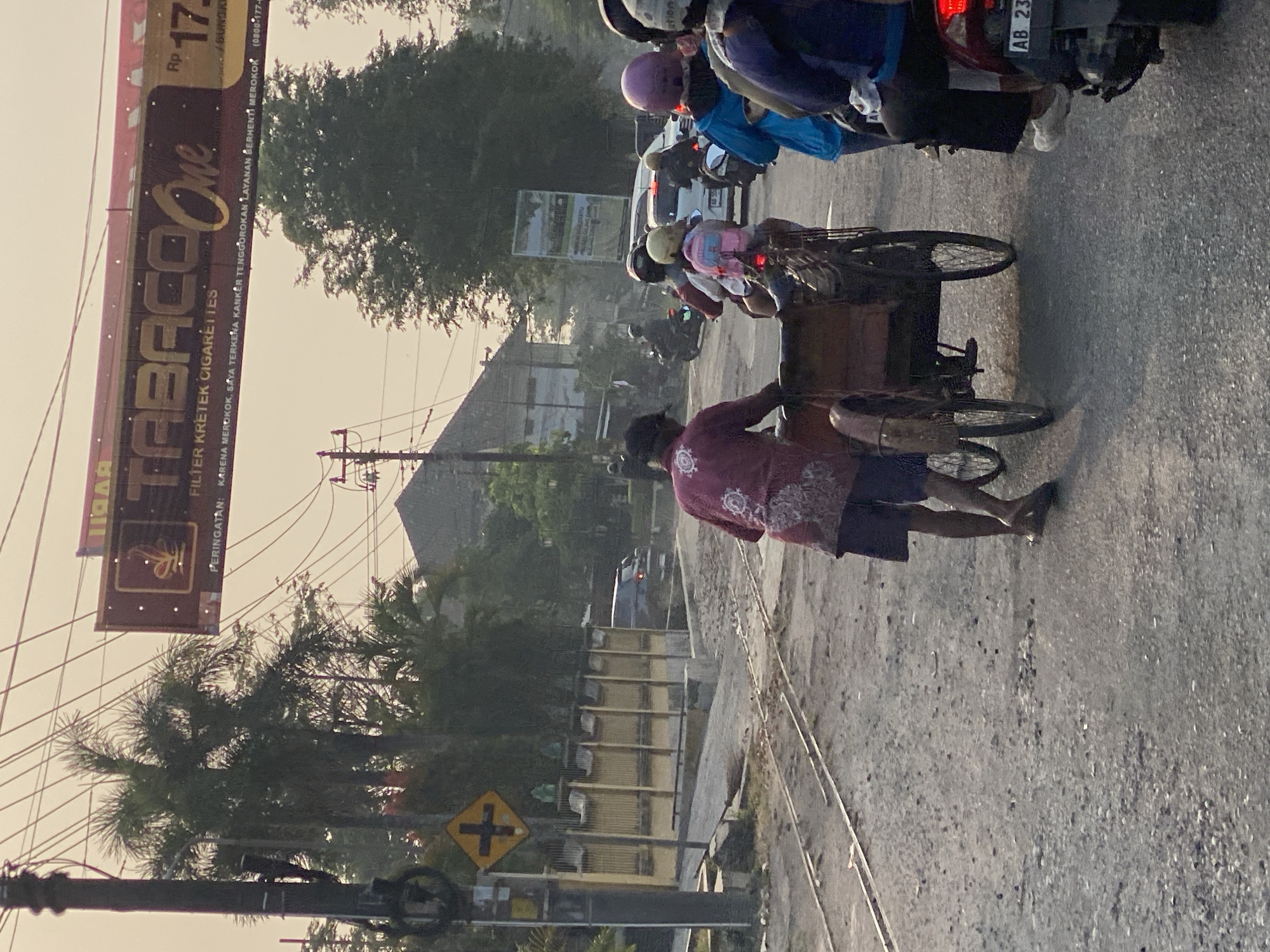 In the city of Kasihan, Indonesia, a street scene unfolds with a man pushing a pedicab. The road is unpaved and appears to be under construction, with loose gravel and visible railway tracks. Overhead, power lines crisscross the sky, and a large billboard advertising "Tabaco One Filter Kretek Cigarettes" hangs prominently. Several motorbikes, some carrying passengers, are present in the street, suggesting traffic. The general atmosphere is hazy, possibly due to dust or pollution, and the lighting suggests it is either early morning or late afternoon. The man pushing the pedicab is wearing a maroon shirt with a patterned design and dark shorts. His bare feet indicate he is barefoot or wearing very thin footwear. The pedicab itself is a three-wheeled vehicle with a wooden cargo area. The surrounding buildings are simple, low-rise structures, typical of a local Indonesian setting.
