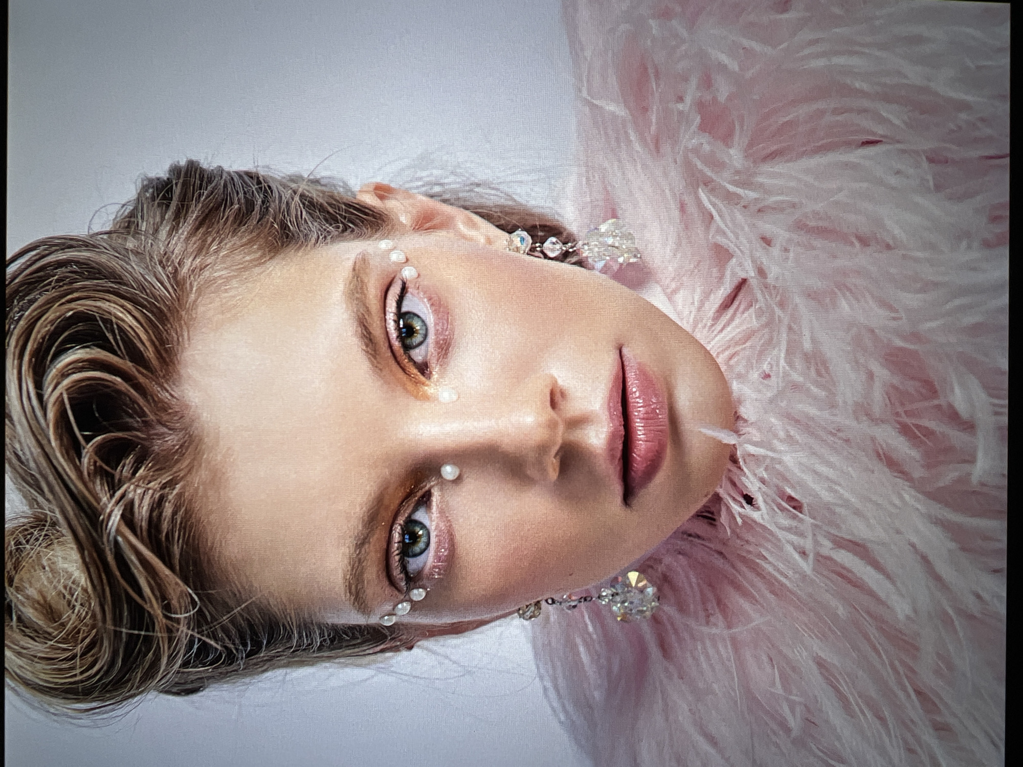 A close-up, head-and-shoulders portrait shows a young woman with her hair styled in a high, swept-back updo. She is wearing elaborate makeup, including shimmering gold eyeshadow and small pearls applied around her eyes, creating an artistic, decorative effect. Dangling crystal earrings adorn her ears, and she is draped in a fluffy pink feather boa or collar that frames her face and shoulders. The background is a plain, light grey. The image appears to be a studio portrait. No location in a city is visible.