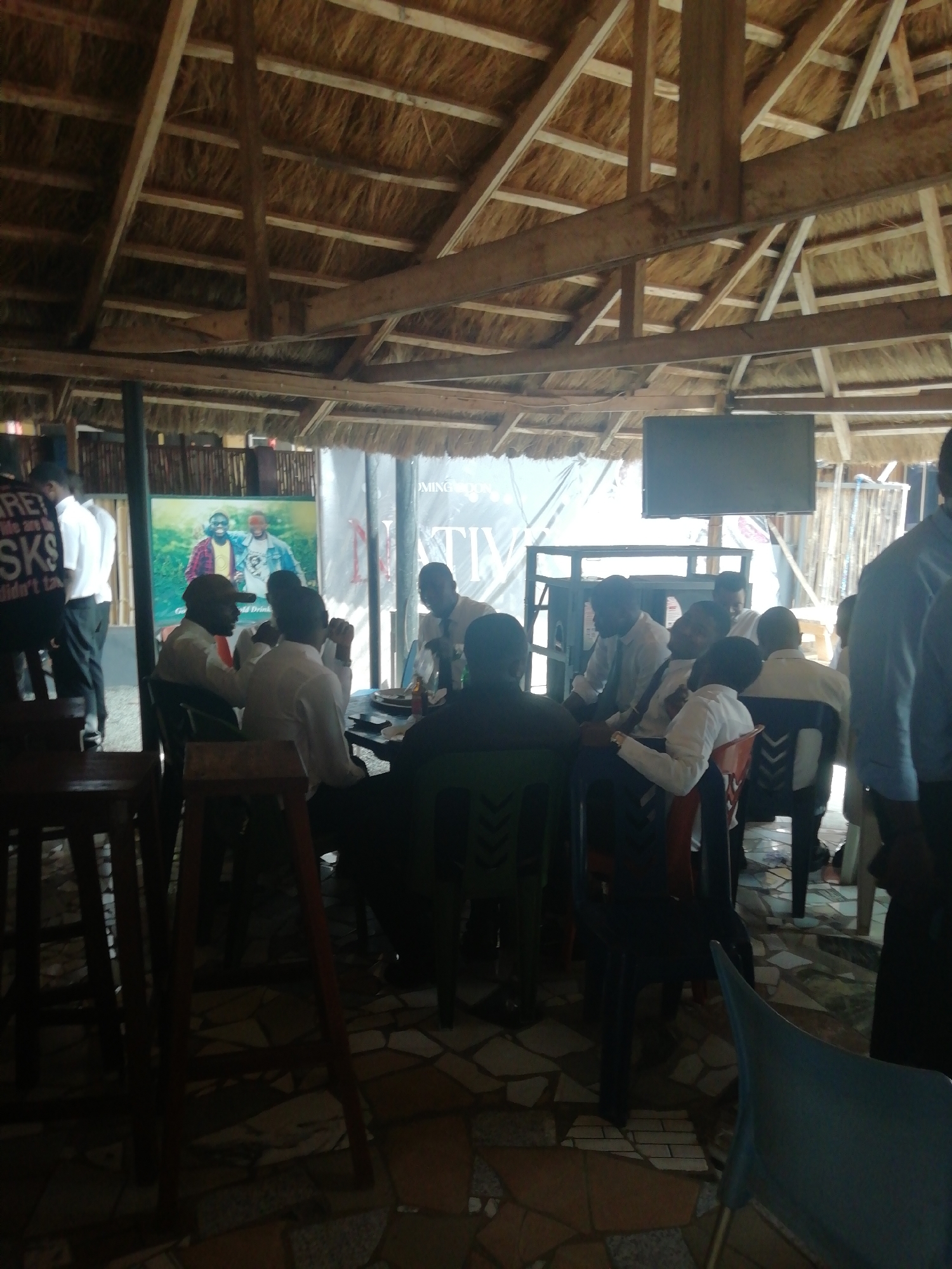 The image depicts an indoor scene in what appears to be a restaurant or bar in Enugu, Nigeria. The roof is thatched, with visible wooden beams supporting the structure. Several men are seated at tables, wearing both casual and formal attire. Some are engaged in conversations, while others appear to be watching a television mounted on a stand. There is a promotional poster visible in the background. The floor is tiled with a mosaic pattern. The atmosphere seems relaxed and social.