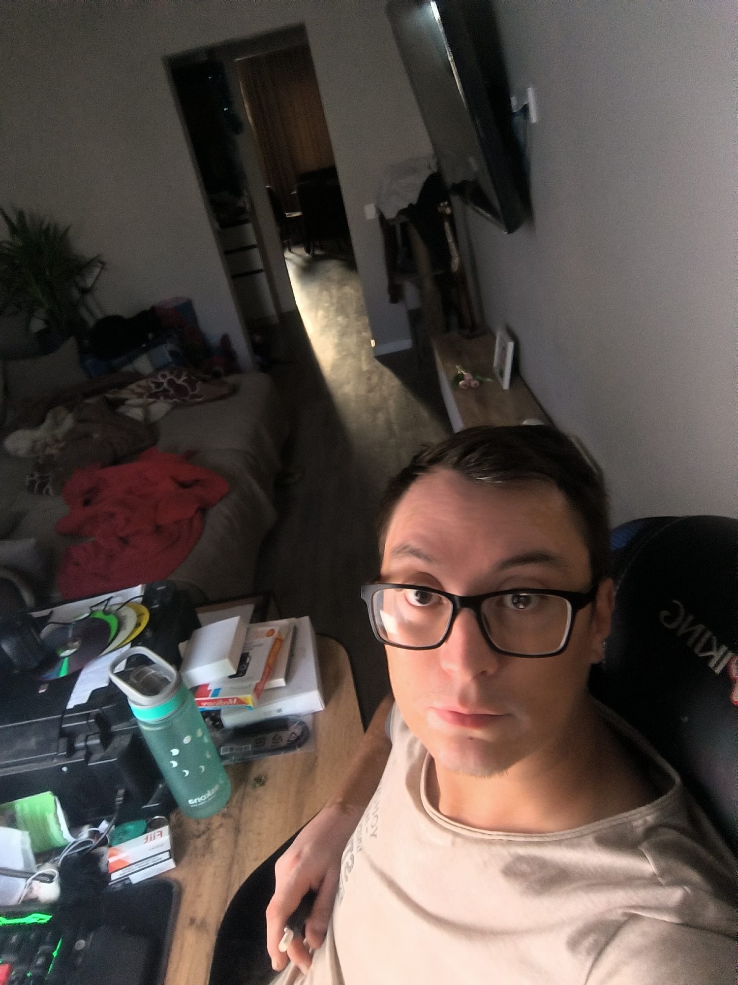 A young adult male with dark hair and glasses is taking a selfie in a dimly lit room. He is wearing a light-colored t-shirt and is seated in a black chair with "VIKING" written on the back. In the background, there is a bed with various items on it, including blankets and stuffed animals. A desk is visible to the left, covered with a water bottle, books, and other objects. A doorway leads to another room, partially obscured by darkness. The overall scene suggests a residential setting. The location is not discernible from the image.