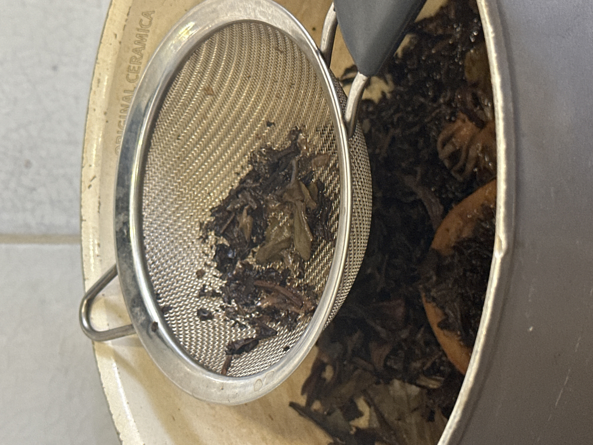 The image shows a close-up, overhead view of a silver mesh strainer filled with dark, loose-leaf tea and some tea leaves in a pot below. The strainer has a metal handle and is resting on the rim of a light-colored pot. The pot's rim has embossed text that appears to read "ORIGINAL CERAMICA". The background is out of focus and appears to be a neutral-colored wall. The lighting is diffused, and there are no people or other objects visible in the frame. The scene appears to be indoors, likely in a kitchen or dining area. The location is Unknown, Unknown.