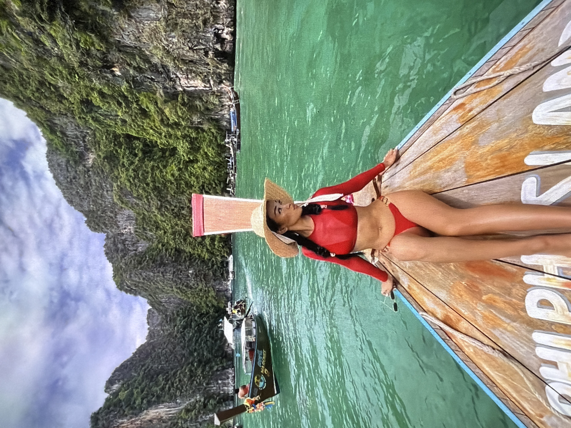A vibrant, sunlit image captures a woman relaxing on a traditional longtail boat amidst a stunning tropical landscape. The woman, with dark hair, wears a red long-sleeved swim top, matching bikini bottoms, and a straw hat. She is positioned on the wooden bow of the boat, leaning on one arm and looking upwards.
The boat floats on calm, clear turquoise-green water, which occupies the middle ground of the scene. In the background, towering limestone karsts or cliffs, densely covered with green foliage and moss, rise dramatically towards a partly cloudy blue sky. Several other longtail boats are visible in the distance, navigating the clear waters, suggesting a popular tourist destination.
The time of day is daytime, with bright, natural lighting. The weather appears fair and pleasant, contributing to the serene and leisurely atmosphere. The visible text on the side of the wooden boat's bow clearly shows "PHIPHI" in white lettering, strongly indicating the location is likely the Phi Phi Islands in Thailand. The overall scene conveys a sense of tropical vacation, relaxation, and natural beauty.