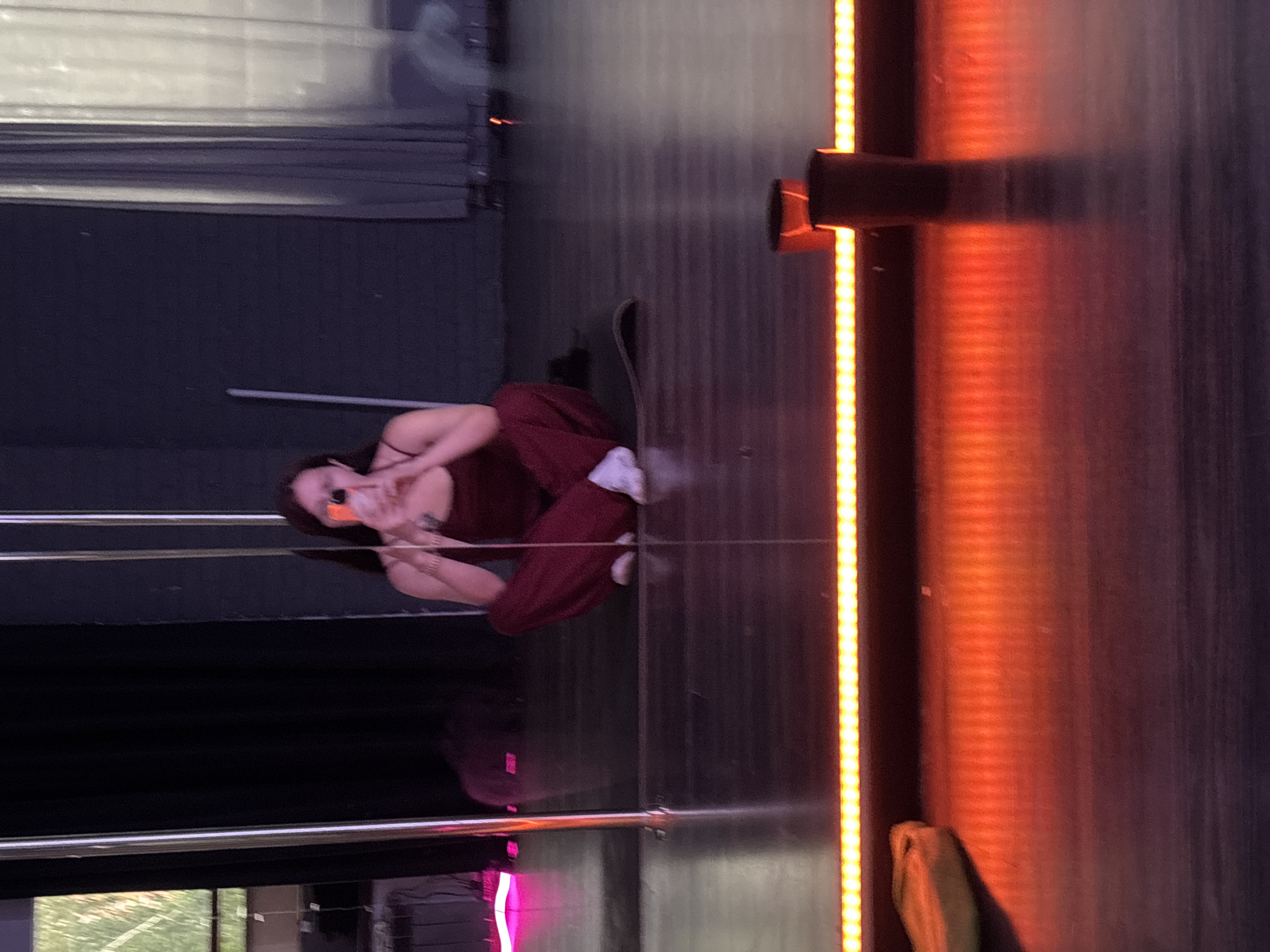 In this vertical, medium shot, a young woman is sitting on the floor with her legs crossed, wearing a maroon outfit and white sneakers. Her head is tilted back, and she's wearing sunglasses. She appears to be taking a selfie with her phone. The background is dark and dimly lit, with a vertical strip of orange lights on the right side of the frame. A metal pole is visible behind her, and a skateboard lies on the floor near her. The scene appears to be indoors, possibly a studio or performance space, given the lighting and the presence of stage-like elements. The mood seems relaxed and casual, with the woman appearing comfortable and engaged in her activity. There are no discernible signs of weather or time of day, but the indoor setting and artificial lighting suggest it could be any time. The location is identified as Naberezhnye Chelny, Russia.