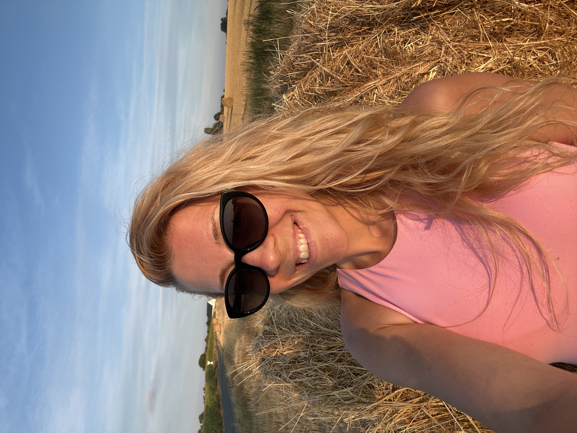 A blonde woman with long, wavy hair and black sunglasses smiles at the camera in a selfie. She is wearing a pink tank top and is positioned in front of a large hay bale. In the background, a rural landscape with a field of wheat, more hay bales, a road, and some houses can be seen under a blue sky with scattered clouds. The lighting suggests it is late afternoon or early evening. The location is identified as Trouville-Alliquerville, France.