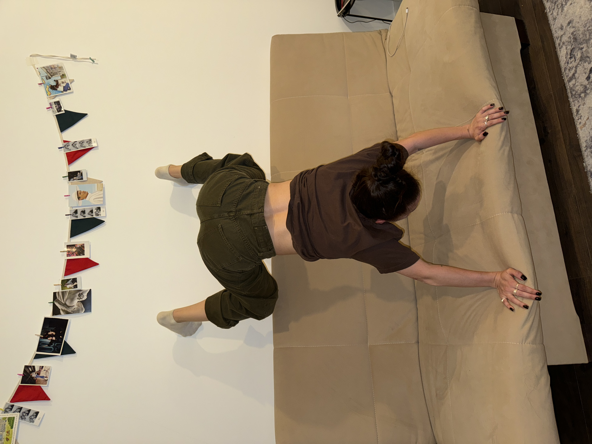 A woman is reclining on a tan sofa, positioned diagonally from the top left to the bottom right of the frame. Her body is oriented with her head near the top right corner and her legs extending toward the top left. She is wearing a brown t-shirt and olive green pants, and her hair is pulled back into a bun. Her arms are stretched out on the sofa, and she is wearing rings on her fingers with dark nail polish.
To the left of the woman, a string of photos and flags is hung along a white wall.
The scene appears to be indoors.
There is no definitive indication of the location being Moscow, Russia.