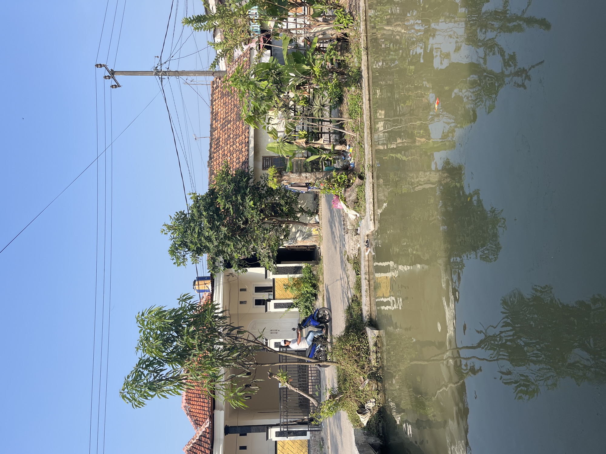 A man rides a blue motorcycle on a paved road next to a body of murky water. The road is lined with houses with red tile roofs and fences. Trees and plants grow along the edge of the water and in front of the houses. Power lines and a utility pole are visible in the background against a clear blue sky. The scene appears to be in a residential area of Laban, Indonesia, as indicated by the architectural style and lush vegetation.