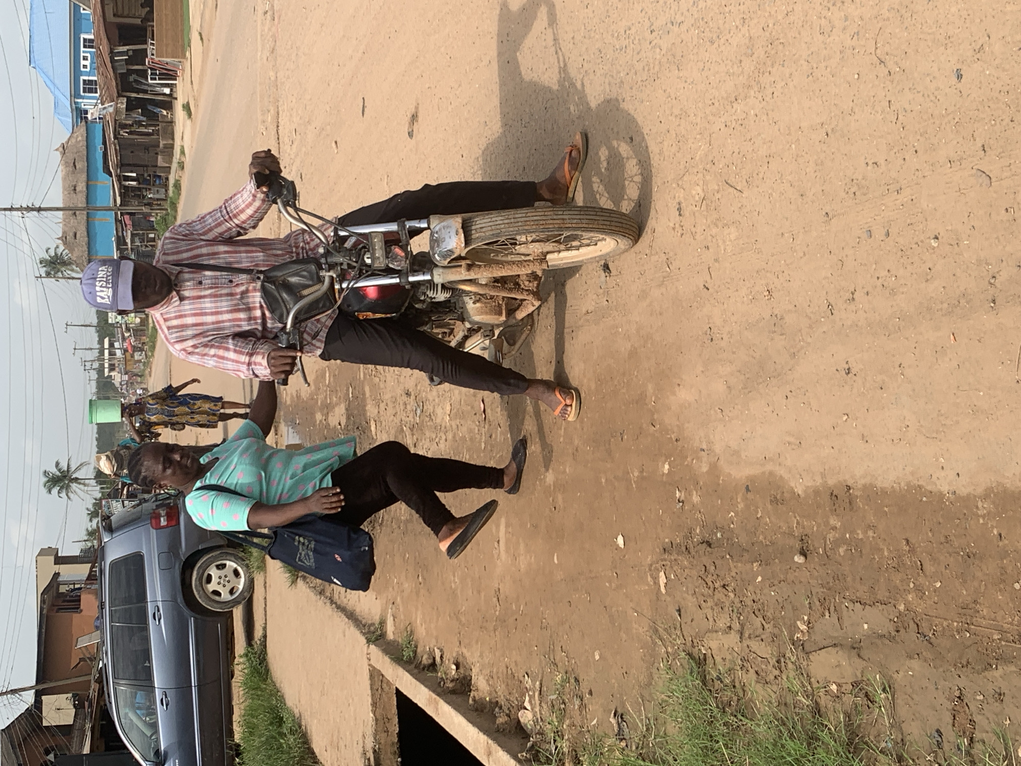 In Shagamu, Nigeria, a man wearing a red and white checkered shirt, black pants, and a purple baseball cap labeled "KATSINA State," drives a motorcycle. He also wears brown flip-flops. A woman with a teal shirt with pink polka dots and black pants is walking with him, carrying a blue bag. Both the motorcycle driver and the woman are dark-skinned.
The motorcycle has a black pouch attached near the handlebars. A blue minivan is parked on the side of the road. Buildings and people are visible in the background. The road is a mix of asphalt and dirt, showing signs of disrepair with puddles. Grass grows along the side of the road. The lighting suggests it is daytime.
