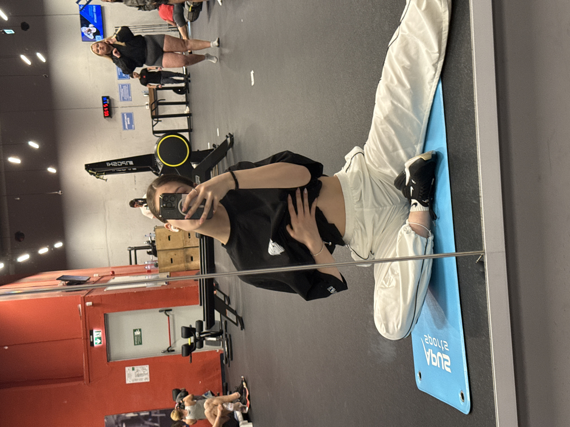 The image captures a mirror selfie taken by a young woman in what appears to be a gym or fitness center. The primary subject is positioned on the right side of the frame, kneeling on a blue exercise mat, with her reflection facing the camera. She is holding a smartphone to take the photo, partially obscuring her face. She wears a black t-shirt, white baggy pants with black trim, and black sneakers with white soles. Her left hand is placed on her stomach.
In the background, the gym environment is visible, characterized by dark grey flooring and industrial lighting fixtures on the ceiling. Various pieces of exercise equipment are scattered around. Directly behind the main subject's reflection, a black rowing machine with "TECHNOGYM" and "SKILLROW" branding is clearly seen. Further in the background, another woman wearing a black top and grey shorts is visible, standing near some equipment. Another person is partially visible near a red wall in the distance. A rack with weights is also present.
Notable details include a digital clock displaying "08:15" on a blue background. Several signs are visible: one in Cyrillic, "СТАТЕГ. УРОК" (Strategy Lesson), and another small blue sign that reads "ТАРИФИ И ЦЕНИ" (Tariffs and Prices). On the blue mat the subject is kneeling on, the words "AQUA SPORTS" are visible in reverse. A red wall or structure with a white emergency exit door, marked with a green 'EXIT' sign and the running man symbol, is also part of the background. The scene suggests an indoor fitness facility, possibly located in Sofia, Bulgaria, given the Cyrillic text.