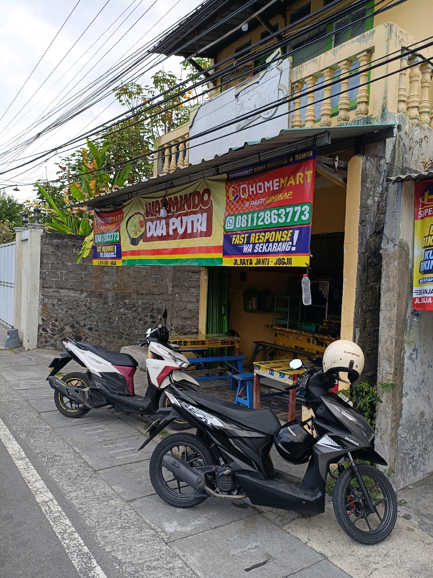 The image depicts a street scene in Purwomartani, Indonesia, featuring a building with a yellow facade and a balcony with decorative pillars. In front of the building, two motorcycles are parked on a sidewalk. The motorcycles are a white and red scooter on the left and a black scooter on the right, which has two helmets attached to it. Above the entrance of the building, there are two banners. The left banner is yellow and red with text that appears to be in Indonesian, and the right banner is red and green with "QHOMEMART" prominently displayed, along with a phone number and address. The building's facade shows signs of wear and tear. Overhead, a dense network of electrical wires crisscrosses the sky. The environment suggests a commercial area, possibly with shops or businesses operating from the building.