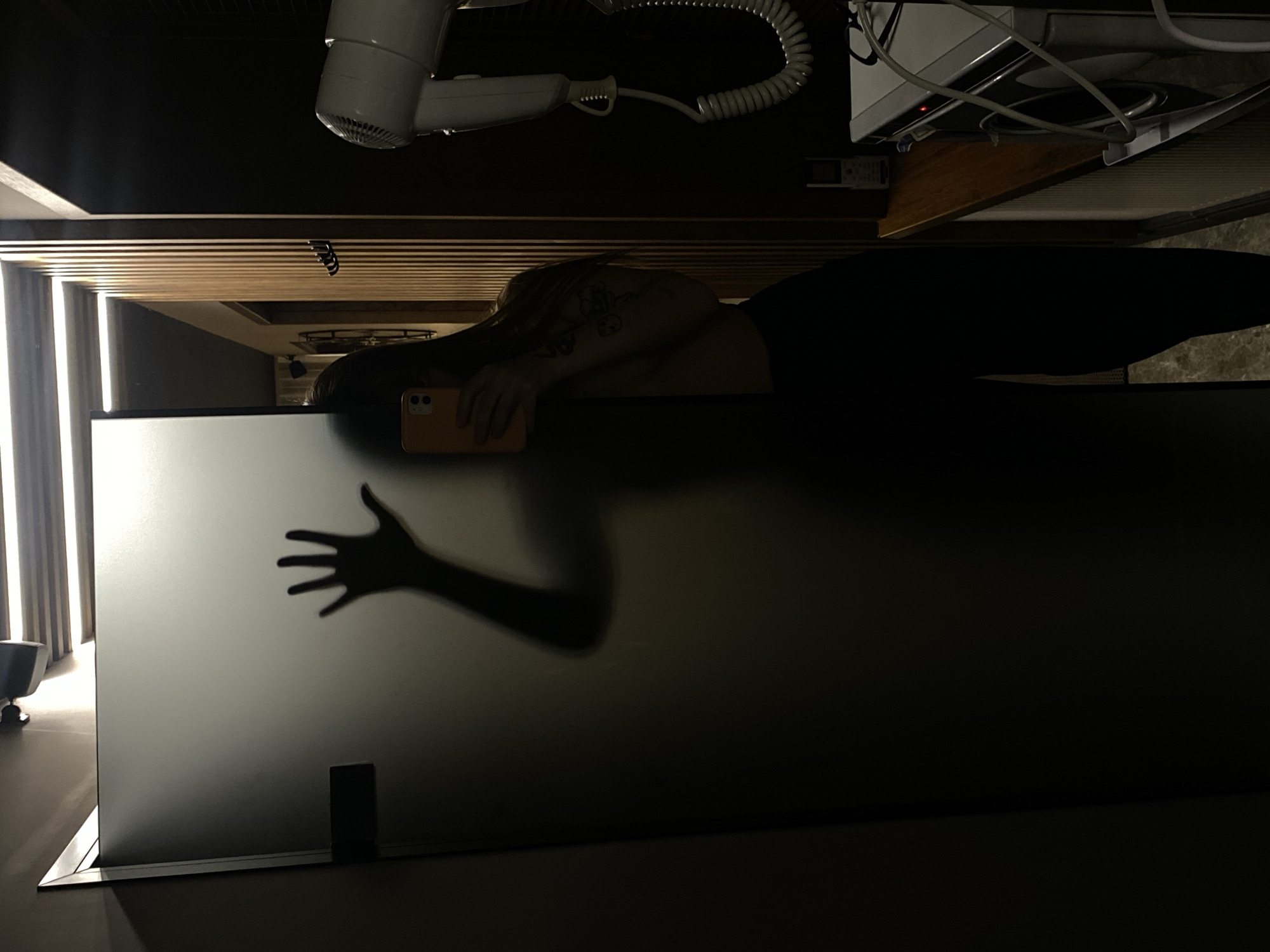 A flipped, high-angle shot reveals a person reclining, holding a cell phone. The person's hair is light, and a tattoo is visible on their arm. They are wearing dark clothing. The person's arm extends, casting a shadow on a frosted glass partition. A hair dryer is mounted on a wall. The location appears to be a bathroom. There is no indication of the city of Votkinsk, Russia.