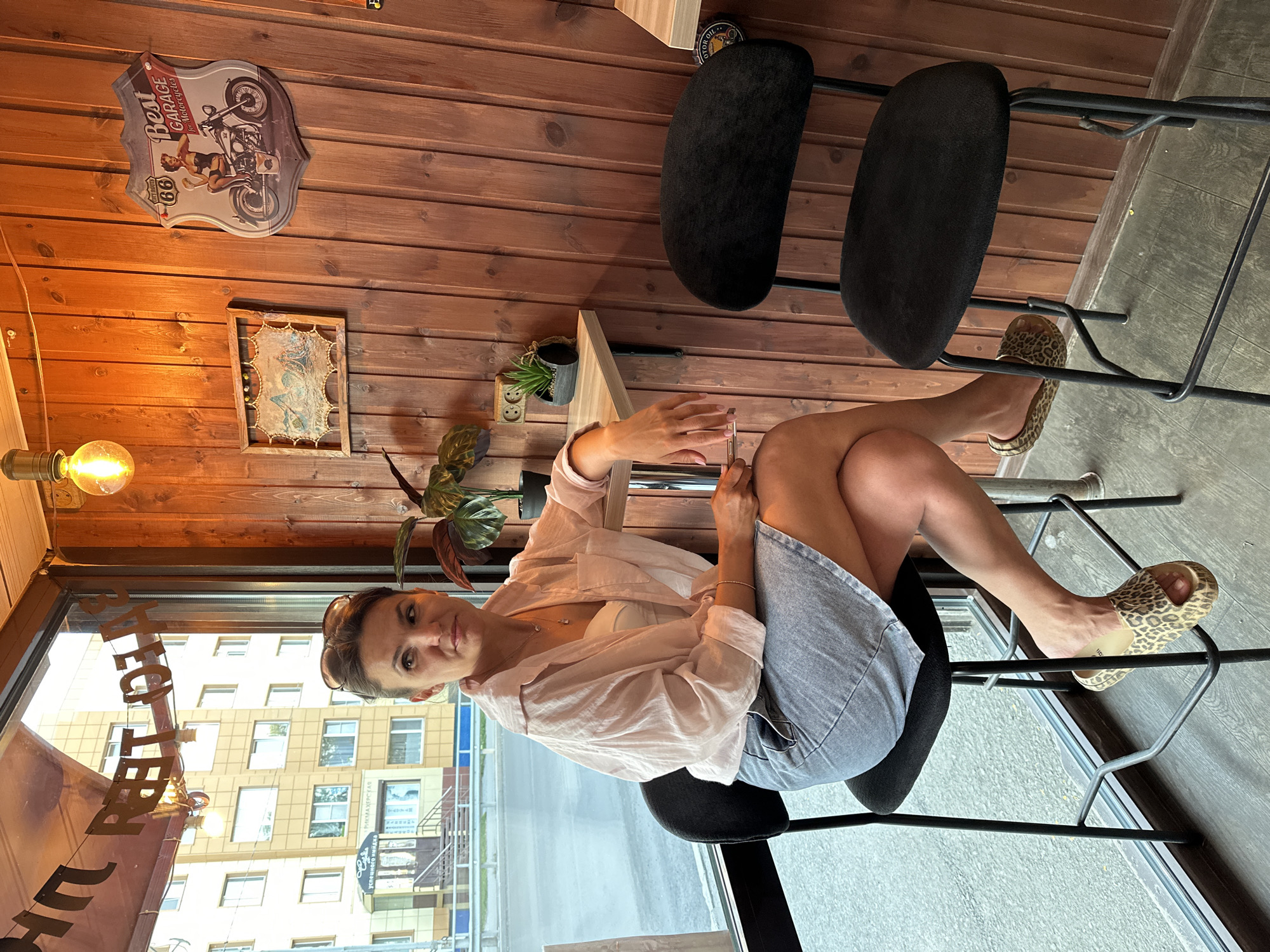 The image depicts a woman sitting on a high stool in what appears to be a cafe or a bar with wood-paneled walls. She is wearing a light pink, open-collared shirt over a white top, and a light blue denim skirt. She has sunglasses on her head and is looking to her left, holding a mobile phone in her right hand. Her legs are crossed. In the background, through a large window, a street with a yellow building and traffic can be seen. The cafe has a vintage or retro theme, with decorative items like a Route 66 sign and a framed picture on the wall. A pendant light bulb hangs from the ceiling. The floor is a grey-green wooden or tiled surface. The overall impression is an indoor commercial establishment with a view of the urban environment. The specific location in Petropavlovsk-Kamchatsky, Russia, cannot be determined from the visual information alone; however, the presence of Cyrillic lettering on a sign outside the window, visible as "Успешного Мира," suggests a Russian-speaking locale.