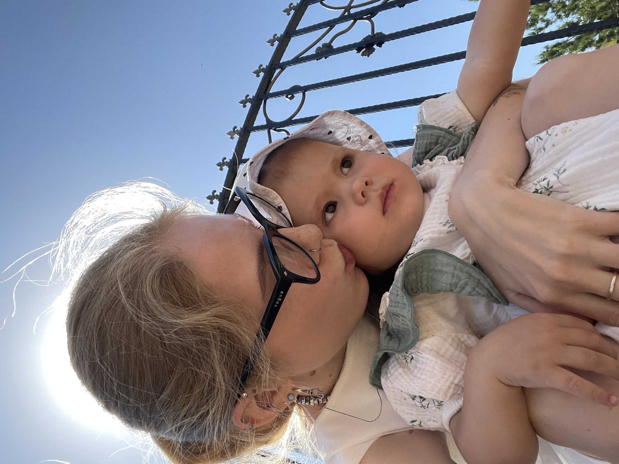The image captures a close-up, low-angle shot of a woman holding a young child. The woman, wearing glasses, appears to be kissing the child on the cheek. The child is dressed in light-colored clothing with a bonnet. Behind them, there is a wrought-iron structure. The background suggests an outdoor setting with a clear, bright sky and intense sunlight. There's no information in the image to suggest it's in Izhevsk, Russia.