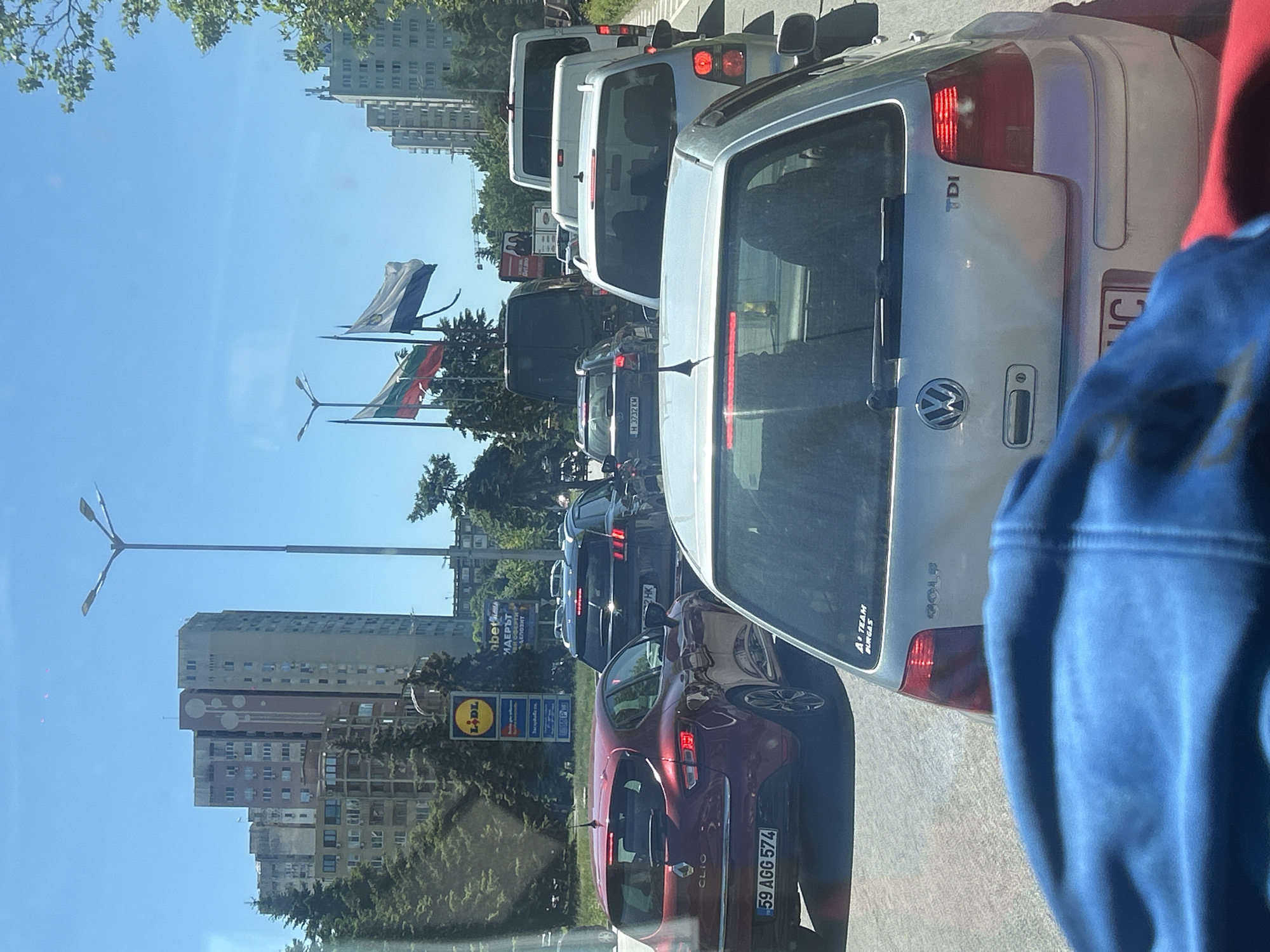 The image shows a street scene in Burgas, Bulgaria, with heavy traffic. The view is likely taken from inside a car, looking at the cars ahead. The vehicles include a red Renault Clio with license plate "59 AGG 574", a silver VW Golf with "TDI" badge and license plate ending in "C", and several white vans. In the background, there are high-rise buildings, trees, and light poles. A Lidl supermarket sign is visible, as are Bulgarian and other flags. A sticker on the Golf says "A Team Burgas". Another car is visible with license plate "H 3732 KM".
