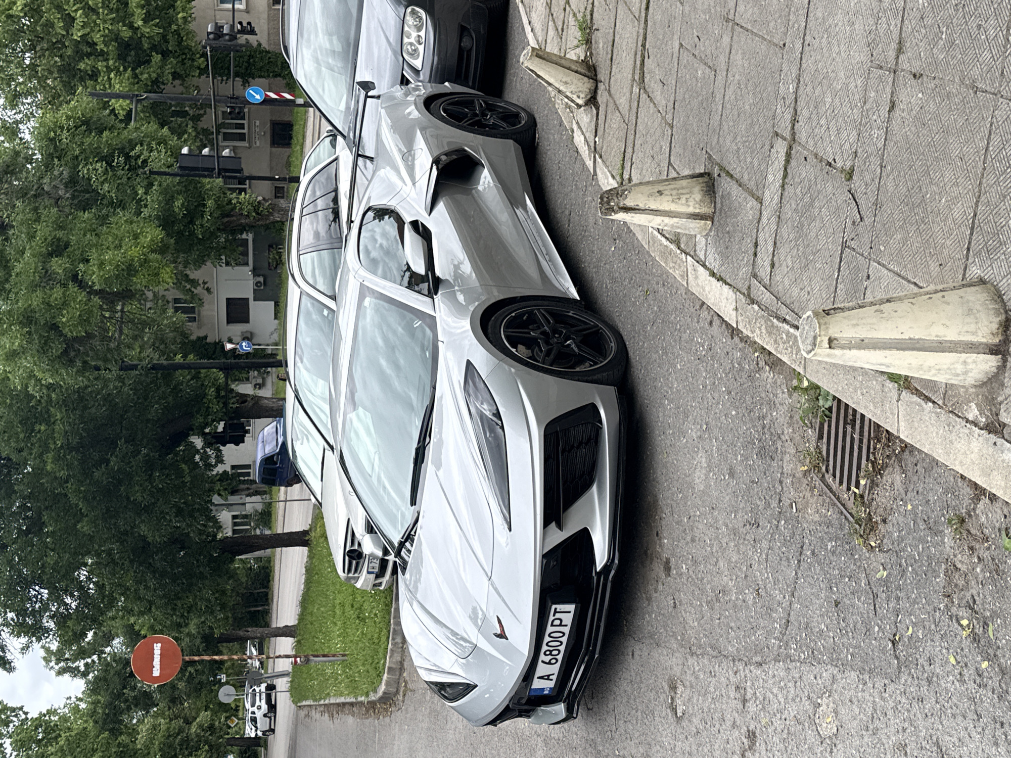 An outdoor, eye-level photograph, rotated 90 degrees counter-clockwise, captures a silver Chevrolet Corvette C8 parked on an asphalt street in Shumen, Bulgaria. The sports car is positioned next to a textured concrete sidewalk, with its front facing towards the viewer's right. The Bulgarian license plate "A 6800 PT" is clearly visible on the front bumper, with 'A' indicating the Shumen region.
Behind the Corvette, several other cars are parked in a line, including a silver or white sedan whose partial grille is visible, and a darker vehicle further back. The sidewalk features several concrete bollards or barriers, some of which appear worn or damaged, and a metal drain grate is visible near the front of the Corvette.
The background is rich with greenery, primarily lush trees, suggesting a leafy urban environment. Buildings are partially visible through and above the tree line. Several street signs and traffic signals dot the scene: a red circular sign with white text displaying "ШУМЕН" (Shumen) is prominent on the left side of the road. Further back, a blue circular sign with a white arrow pointing right is seen near an intersection with traffic lights. Another smaller blue directional arrow sign is also visible.
The scene is lit by soft, diffused daylight, indicating an overcast or cloudy sky, as there are no harsh shadows. The overall impression is one of a typical daytime street scene in an urban area, characterized by parked vehicles and street infrastructure.