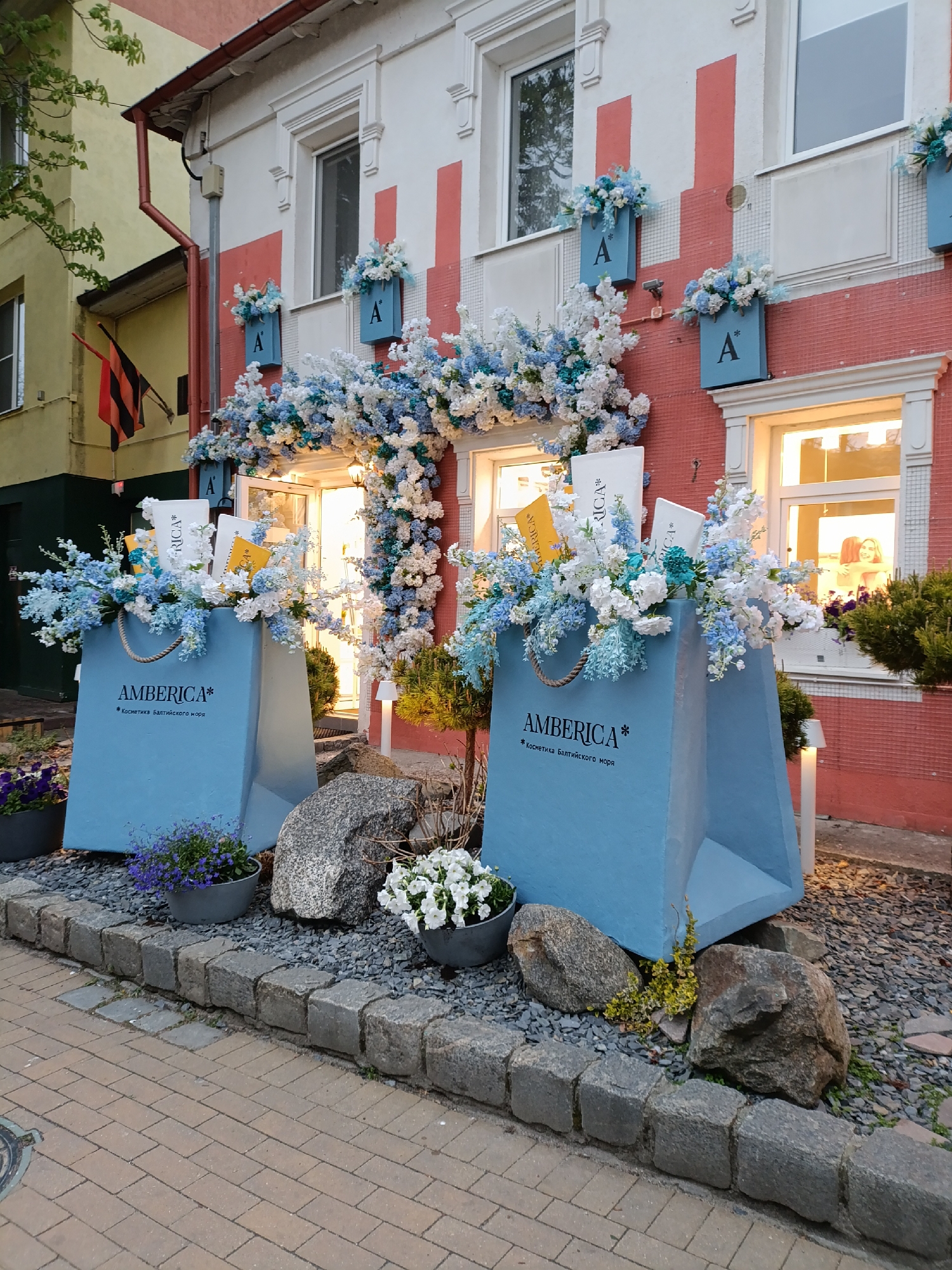 Here's a detailed description of the image:
**Scene:** The image captures the exterior of a shop in Zelenogradsk, Russia. The shop, likely a cosmetics store named "AMBERICA*", is decorated with blue and white floral arrangements to create a visually appealing storefront.
**Building:** The building has a red façade with white accents. There are windows on the upper level with flower boxes underneath, each bearing the letter "A". The shop's entrance is adorned with a floral arch.
**Shopfront Display:** Two oversized, light blue shopping bags with the store's name and tagline "Косметика Балтийского моря" (*Baltic Sea Cosmetics*) printed on them flank the entrance. Each bag is filled with blue and white flowers, along with product packaging (primarily white with "AMBERICA*" visible, and yellow/gold colored).
**Ground Level:** The base of the displays is a rock garden featuring various sizes of grey stones and plants, including some in grey pots. A border of stone paving separates the display from the brick paved sidewalk.
**Adjacent Building:** Partially visible to the left is a yellow building with a black, orange, and red flag.
**Overall Impression:** The scene presents a well-maintained and thoughtfully decorated storefront, likely intended to attract customers with its aesthetic appeal. The combination of colors, flowers, and the oversized shopping bags creates a memorable and engaging display.