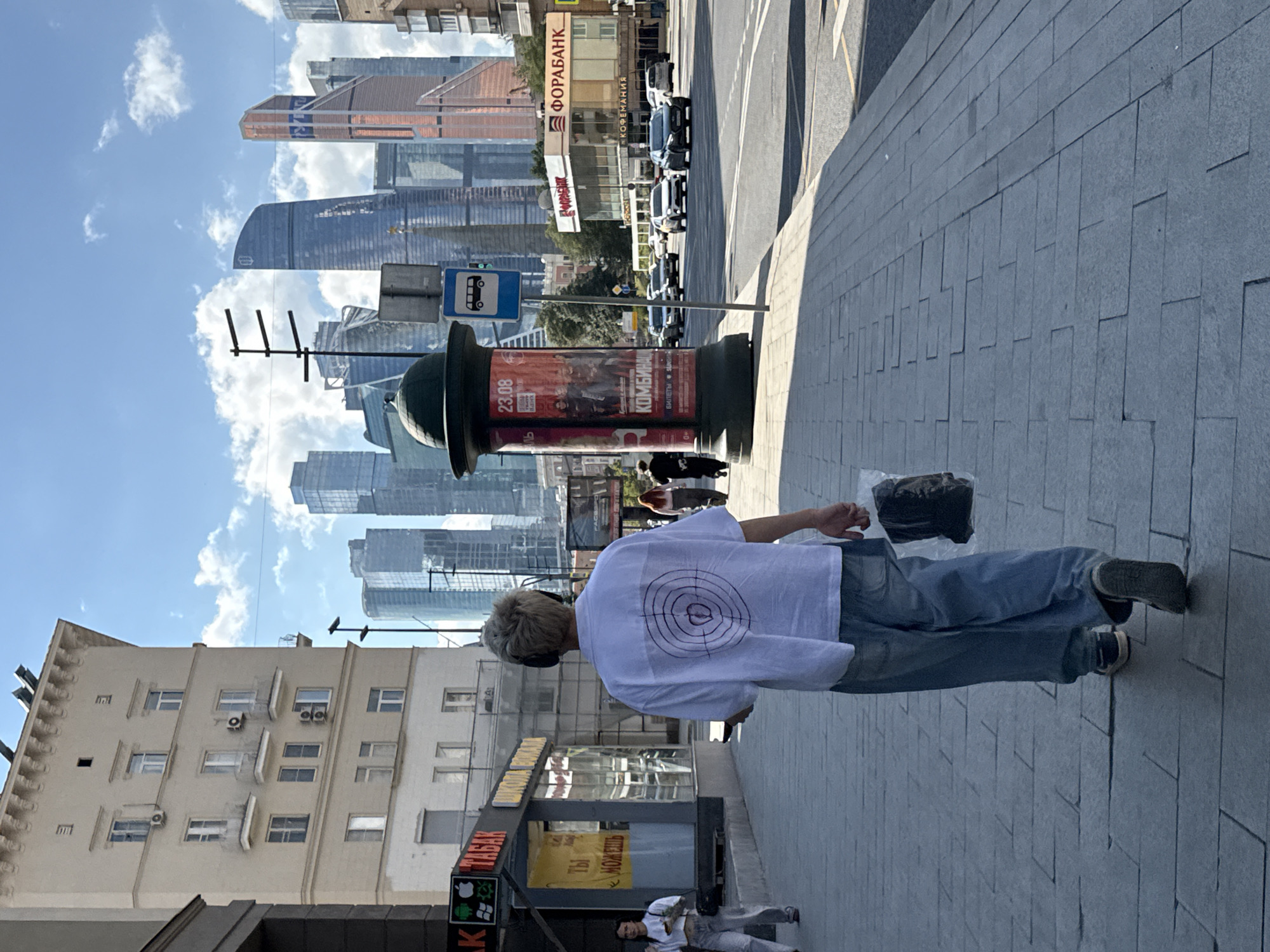 The image shows a street scene in Moscow, Russia, with the Moscow International Business Center in the background. A person wearing a white t-shirt with a dark circular design and blue jeans is walking away from the camera on a paved sidewalk. The person is wearing headphones and carrying a clear plastic bag with a dark item inside. To the left of the person is a kiosk with signs in Russian, including "ТАБАК" (tobacco). In the background, modern skyscrapers dominate the skyline under a bright blue sky with scattered clouds. A bus stop sign is visible on the right side of the street, along with cars driving on the road. The overall scene depicts a typical urban environment in Moscow on a sunny day.