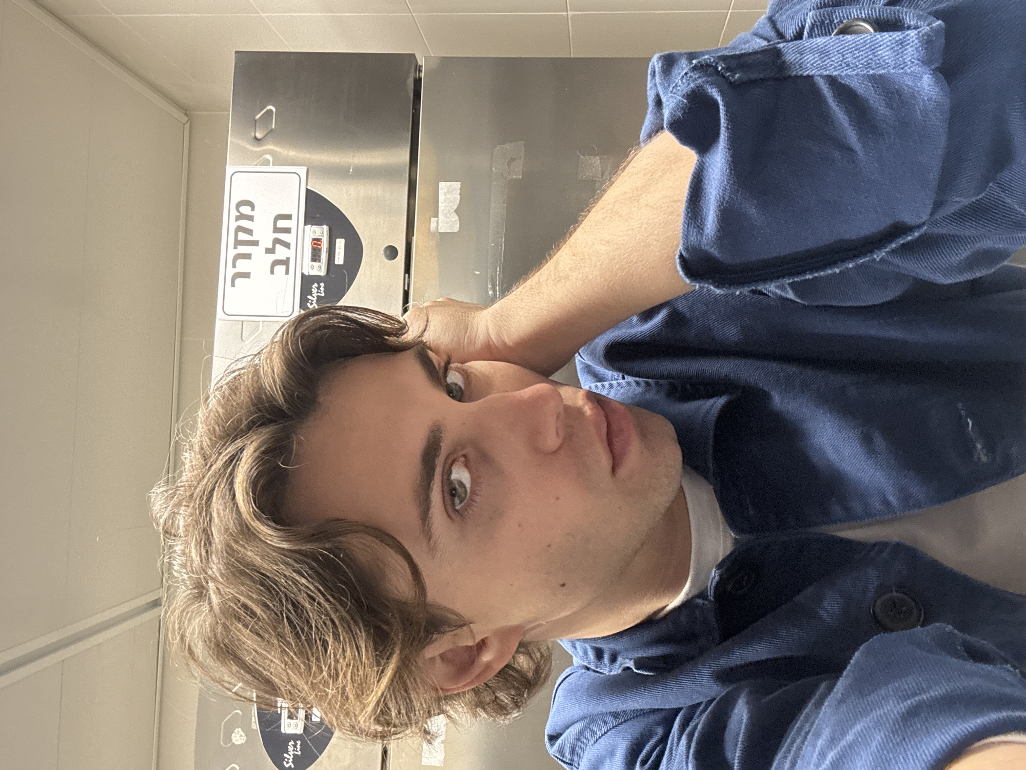 A selfie taken indoors in a commercial setting, potentially a restaurant or cafeteria. The subject is a young man with brown, wavy hair and light eyes, wearing a blue button-up shirt over a white undershirt. He is looking at the camera with a neutral expression, his hand resting on his head.
Behind him is a stainless steel refrigerator or freezer unit with a sign in Hebrew, which translates to "Milk Refrigerator," and the brand "Silver Line." The surrounding environment appears to be a utilitarian space with white walls and possible tile flooring.
The location is likely in Tel Aviv, Israel, given the Hebrew signage.