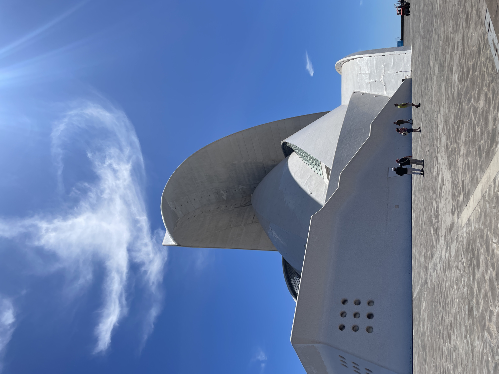The image depicts the Auditorio de Tenerife Adán Martín in Santa Cruz de Tenerife, Spain. The architectural structure features a prominent curved roof and white concrete construction. A group of four people are walking along the walkway near the building. The sky is blue with scattered white clouds and sun glare. The ground surface consists of grey stone pavement. A piece of construction equipment is visible at the top right of the scene.