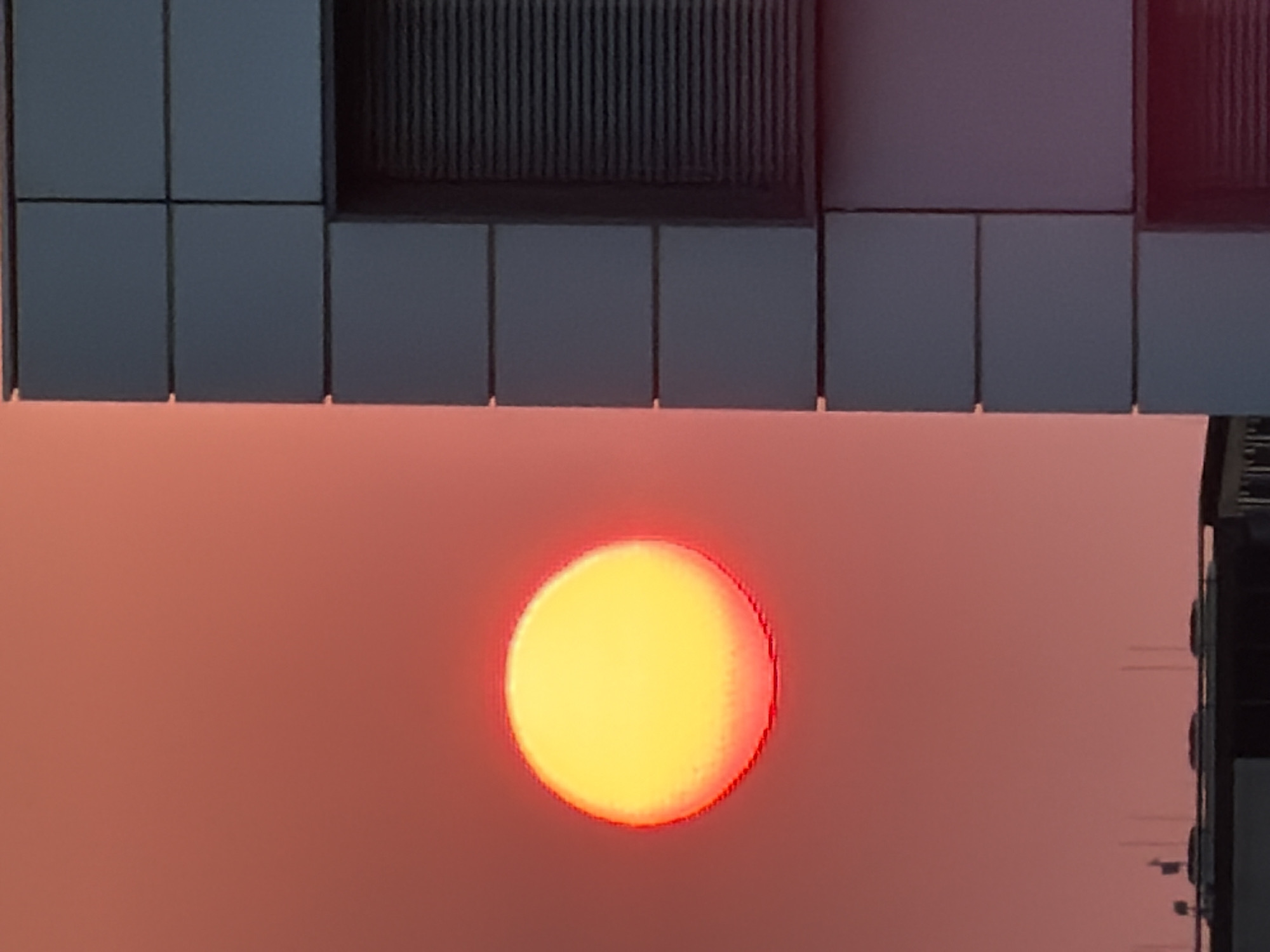 A large, bright yellow sun with a red halo is visible in a hazy reddish-orange sky. To the right of the sun, a section of a modern building with dark gray paneling and horizontal blinds on a window is present. In the lower part of the image, a dark building structure with several antennas on top is partially visible, suggesting an urban or industrial setting. The image captures a sunset or sunrise scene, likely in Leudelange, Luxembourg.