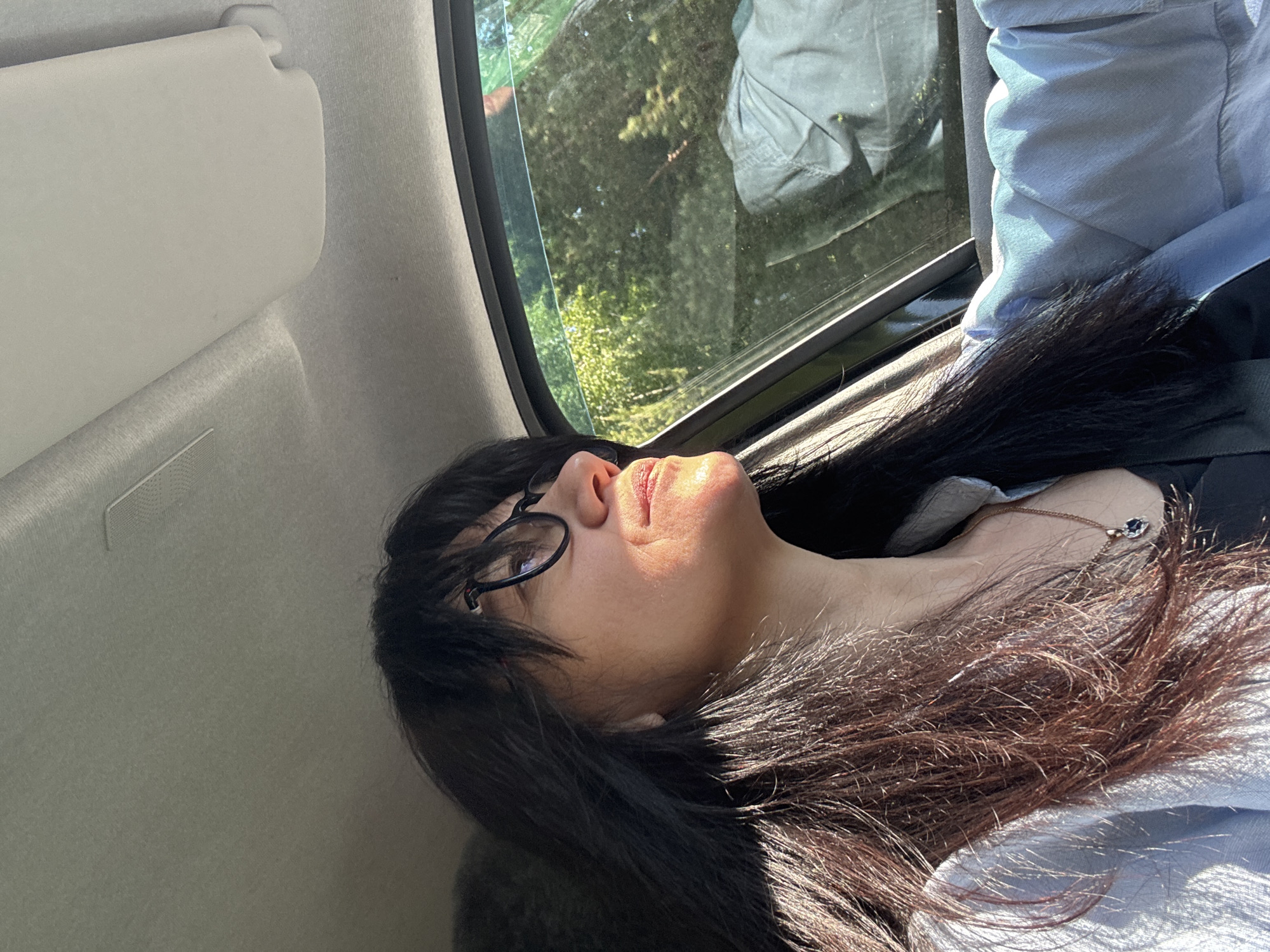 A person is lying down inside a vehicle. They have long dark hair, glasses, and a necklace. The person is facing upwards towards a window. Outside the window, there are trees and another person in the reflection. A sun visor is visible on the left. The location is Chaikovsky, Russia.