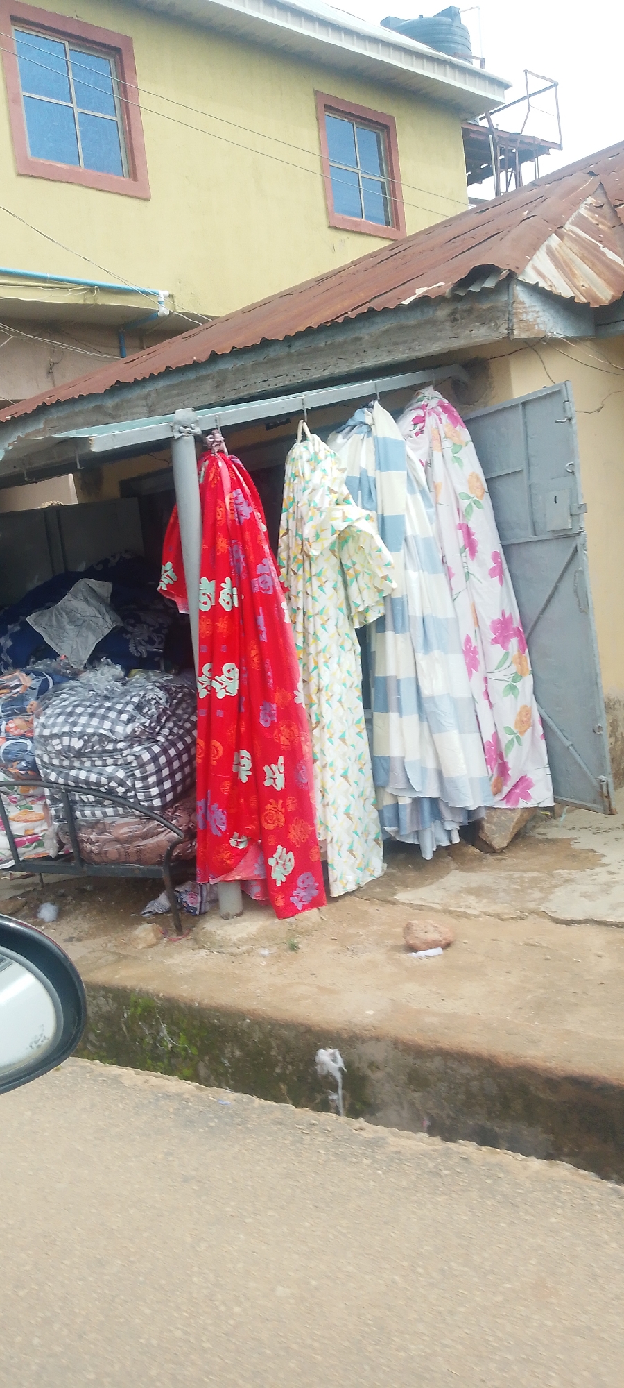 The image shows a storefront in Jos, Nigeria, with fabric items hanging outside. The building is painted yellow with two windows and a rusted metal roof. In the foreground, several draped fabrics are visible, including one red with floral and Chinese character prints, a white one with a geometric pattern, and others with stripes and floral designs. To the left, stacks of fabric or bedding with checkered patterns are piled on a metal rack. A partially visible car side mirror is in the bottom left corner. The ground appears to be a mix of dirt and concrete, with some debris and a drainage ditch. The scene suggests a marketplace or retail stall.