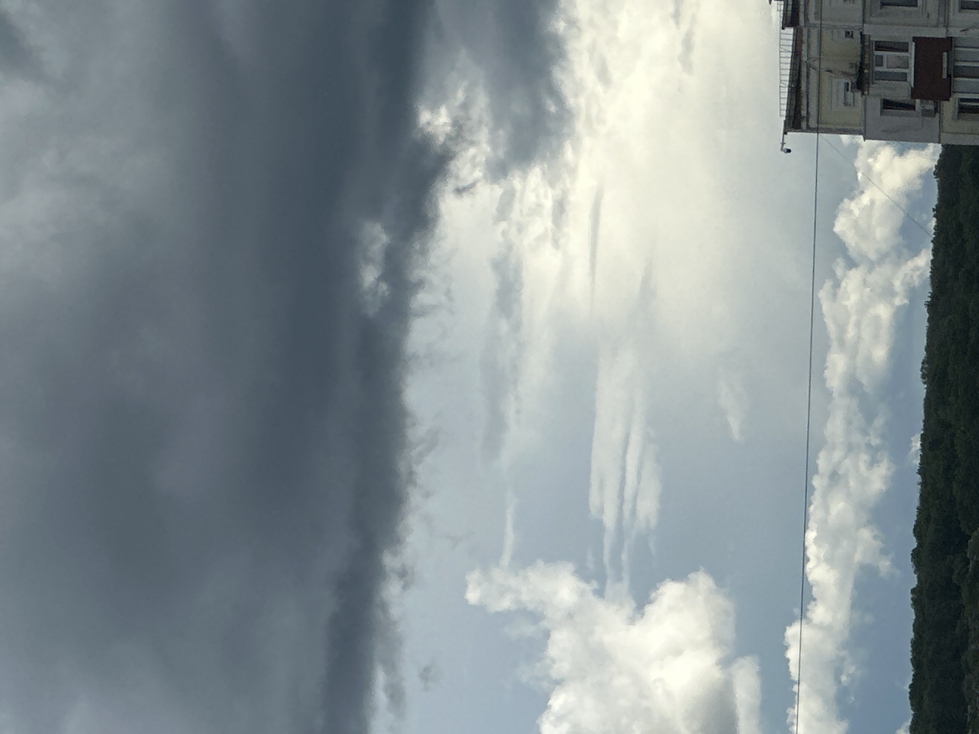The image depicts a cloudy sky with a building on the right side. To the right of the building there is a row of green trees. The sky is a mix of dark gray and bright white clouds, indicating a possible storm or changing weather conditions. An electrical wire runs vertically through the center of the image.