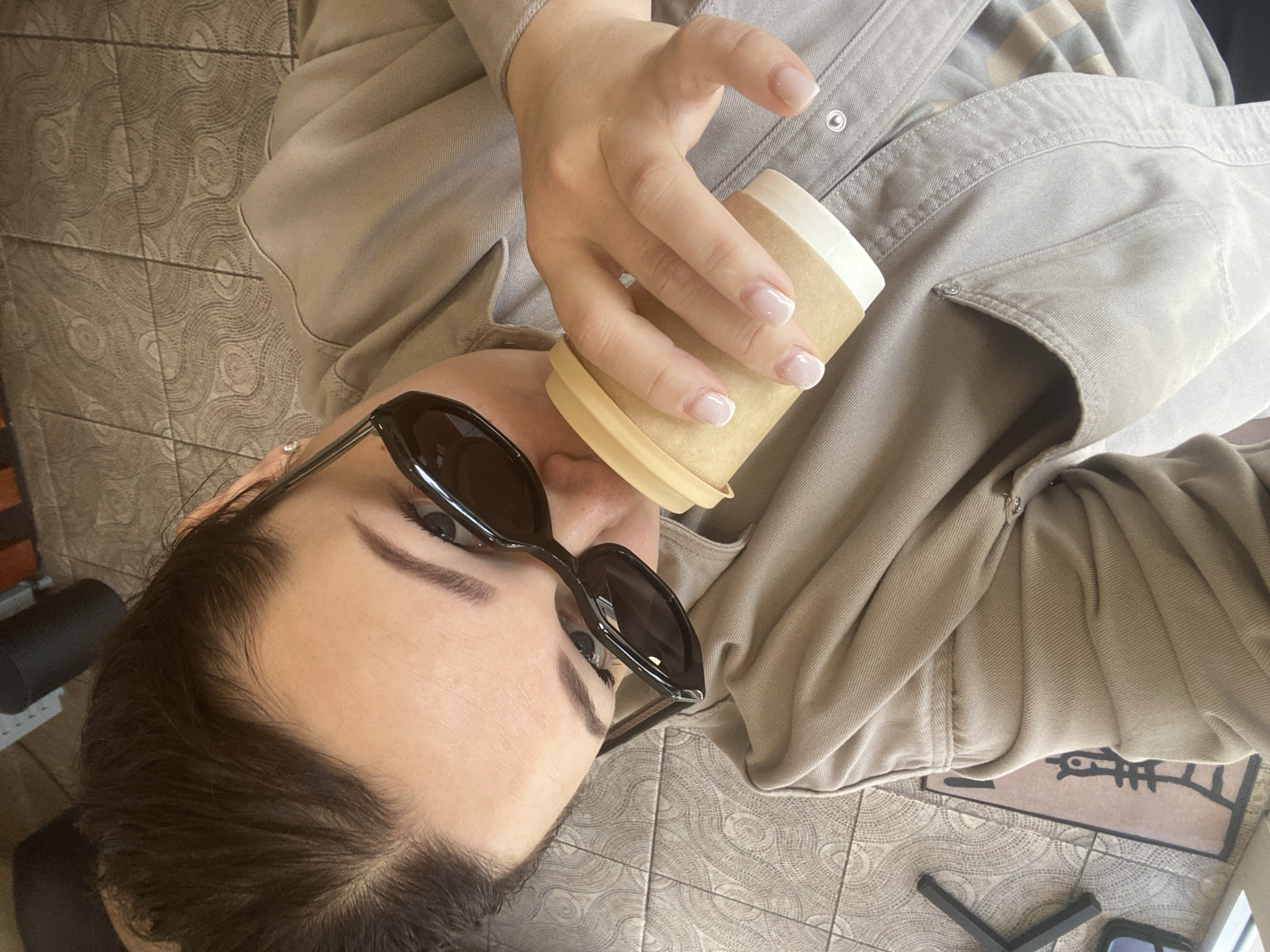 An overhead shot captures a person, likely female, in a casual indoor setting in Sochi, Russia. The individual, with dark hair and neatly groomed eyebrows, wears large dark sunglasses that obscure their eyes. They are seen holding a light brown disposable coffee cup with a beige lid to their mouth, suggesting they are drinking or about to drink from it. Their right hand, visible in the frame, features a neat French manicure.
The person is dressed in a light beige or khaki-colored collared garment, possibly a jacket or shirt, which has visible snap buttons. A hint of a striped or patterned undergarment can be seen beneath.
The background consists primarily of a light brown tiled floor, characterized by a repeating pattern of concentric circles or wavy lines. In the lower right of the frame, a dark doormat is partially visible, featuring a stylized light-colored design resembling a fish skeleton or abstract symbol. A dark, angular object, possibly part of a chair or table leg, is also present on the floor. No discernible text is clearly legible in the image. The overall impression is one of a relaxed, casual moment.