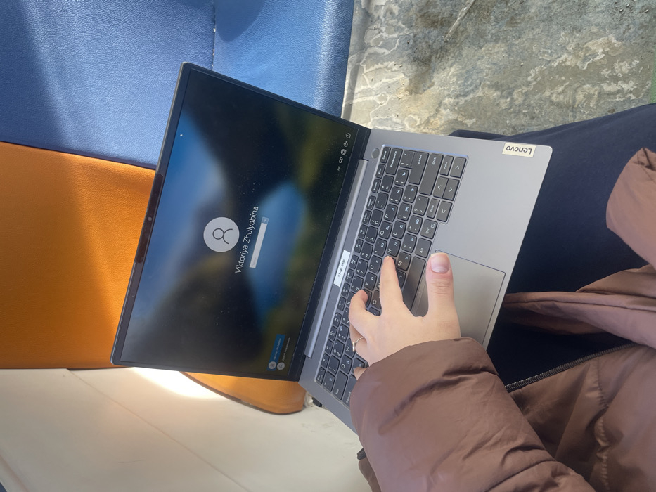 An overhead-angle image captures a person's left hand on a laptop keyboard, resting on their lap. The person wears a brown, puffy jacket and dark blue trousers. The hand, adorned with a ring on the fourth finger, is positioned over the left side of a silver/grey Lenovo laptop's keyboard, which features both Latin and Cyrillic characters. A white sticker with black text "CT-A9E-0440" is visible on the laptop's palm rest. The laptop screen displays a Windows login interface with a blurred landscape background, a user profile icon, and the name "Viktoriya Zhulyabina." Behind the laptop, two sections of worn upholstered seating are visible: one bright orange to the left, and one blue to the right. Bright sunlight illuminates the scene from the left, casting a reflection on the orange seat and laptop screen. The floor or ground surface beneath the person's legs appears light-colored, textured, and shows signs of wear. The specific location within the city of Бабеево, Russia, is not discernible from the visual evidence in the image.