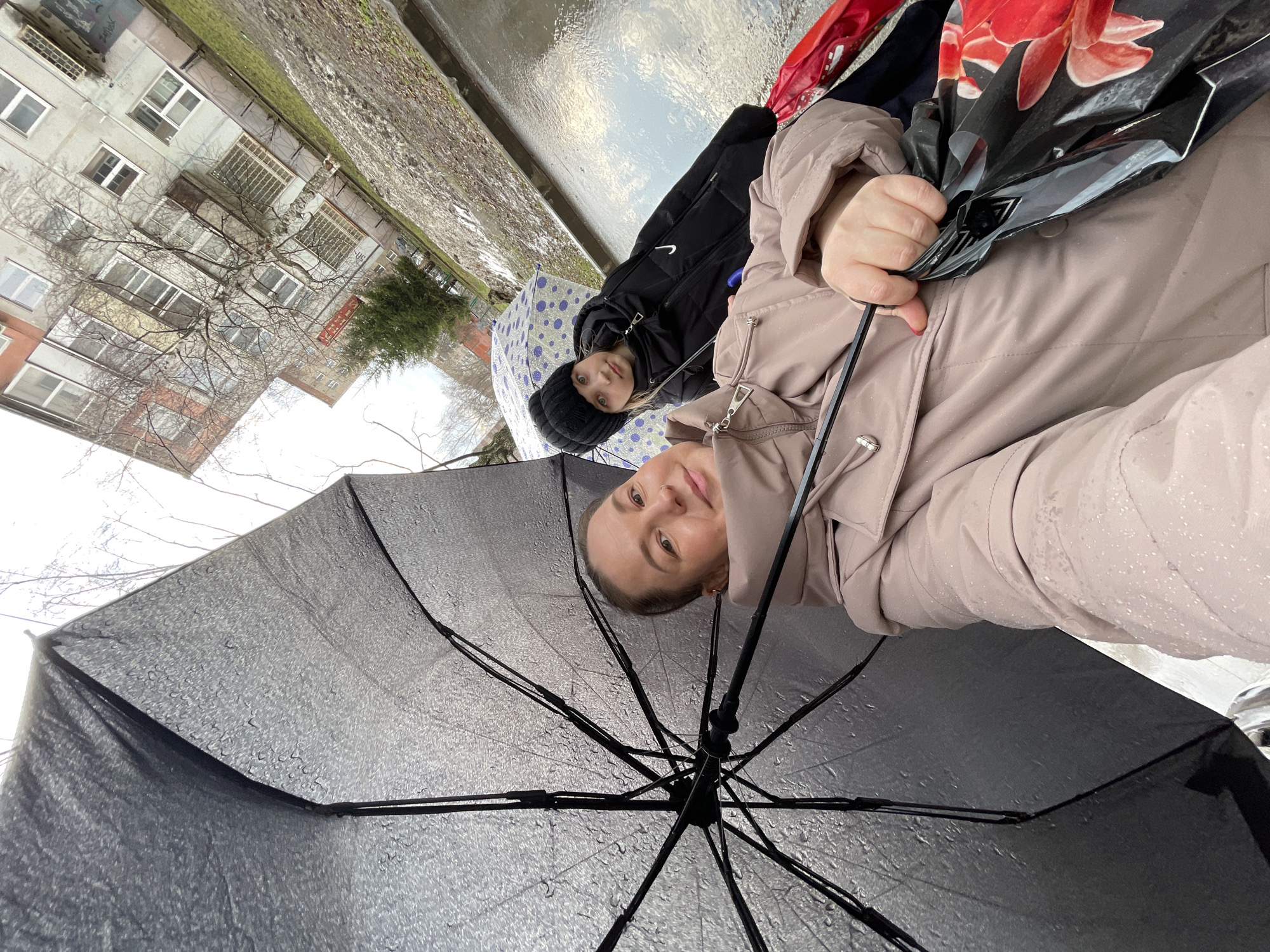 Two individuals are visible in a photograph taken from a low, upward-looking angle, likely a selfie perspective under a black umbrella with raindrops. The person in the foreground is fair-skinned, wearing a light-colored puffer jacket, and looking directly at the camera. The second person, situated slightly behind and to the left, is wearing a black jacket and a dark-colored beanie, and also appears to be looking toward the camera. A blue umbrella with white polka dots is partially visible behind the second person.
The background features an urban setting. There is a wet paved area that possibly indicates a street or sidewalk. The architecture suggests the city of Izhevsk, Russia, with multi-story residential buildings visible. The buildings are of a typical Soviet-era design, characterized by their boxy shapes, rows of windows, and simple facades. A tree with bare branches can also be seen in the background. The photograph was likely taken on a rainy day in Izhevsk.