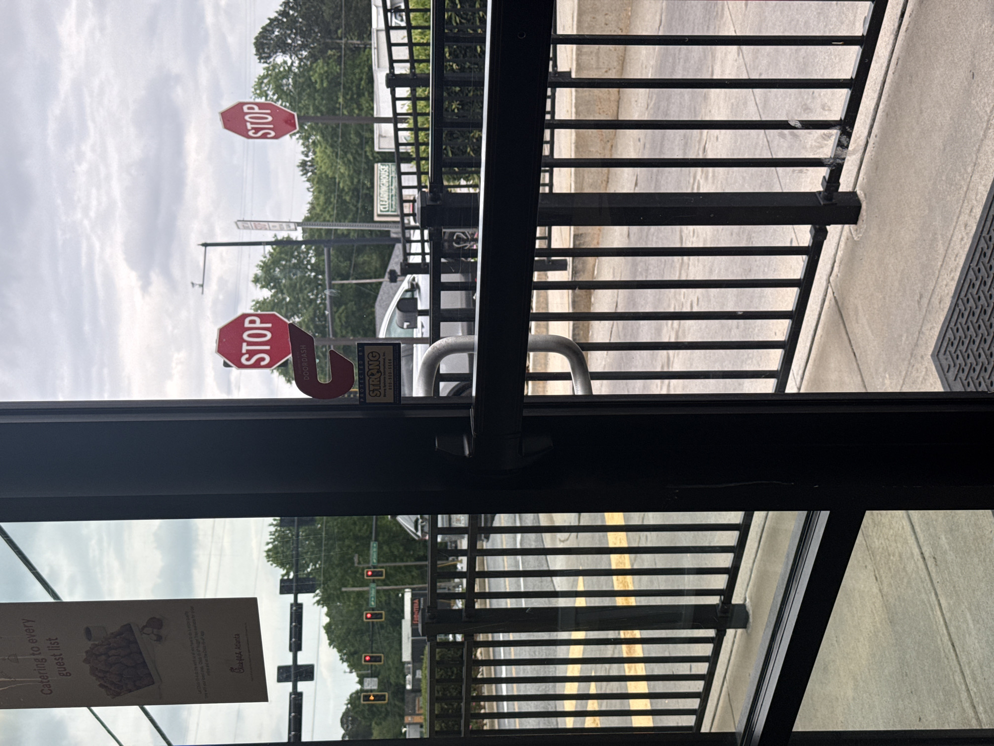 This is an indoor shot from what looks like the inside of a Chick-fil-A restaurant in Stone Mountain, Georgia. Looking out the restaurant door through the metal safety bars, there are stop signs, traffic lights, and street lines painted on the road. The stop signs are partially obscured, with a "Protected by Strong" sign. A Chick-fil-A poster for catering is visible in the left corner. Outside on the street is a Clearinghouse building.