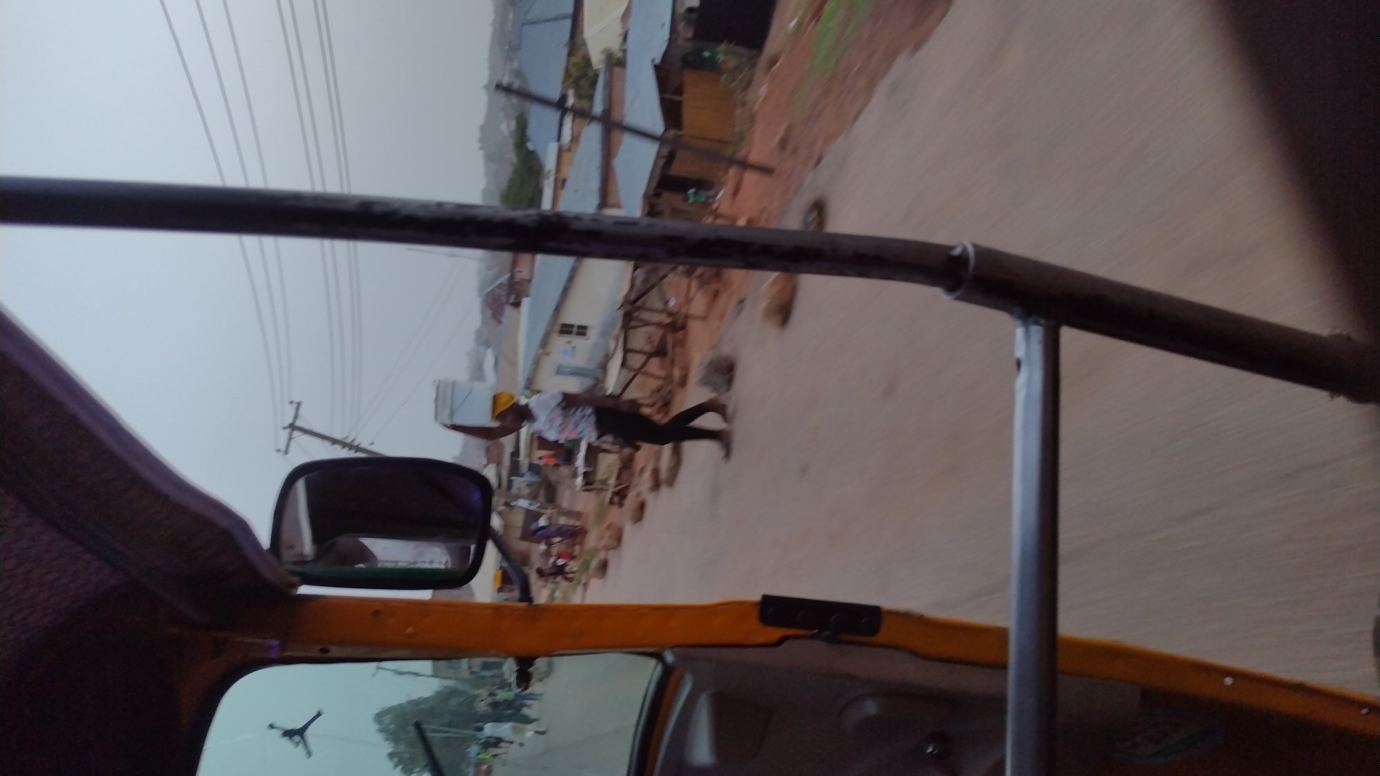 The image appears to have been taken from inside a vehicle, likely a "keke" (tricycle taxi), a common mode of transport in Jos, Nigeria. The view is somewhat obstructed by the frame of the vehicle, including a side mirror and part of the roof. Through the windshield, a street scene unfolds.
A man wearing a yellow hard hat, a patterned short-sleeved shirt, and dark trousers is carrying a white, rectangular object. He is walking on the side of a road past buildings, some of which are constructed of red earth bricks. Power lines stretch above the street and buildings.
The sky is overcast and the light is diffused.
