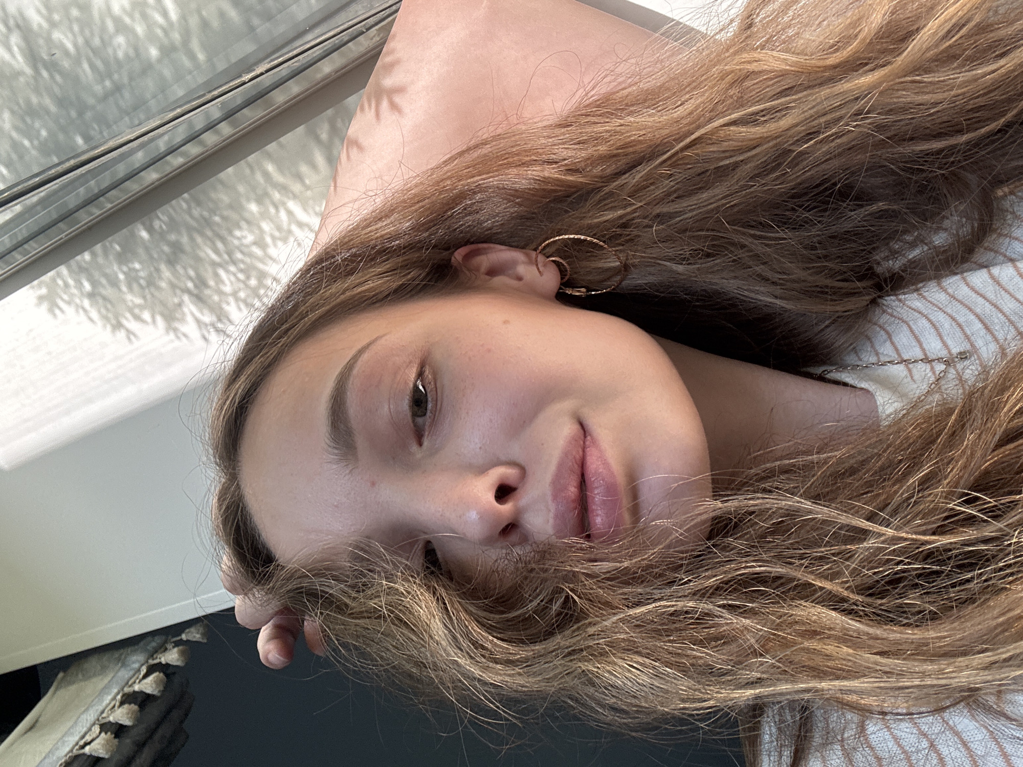 A young woman with long, wavy brown hair is captured in a close-up selfie. She is wearing a striped sweater and a delicate necklace. Her right hand is raised, gently touching her hair near her forehead. She has hoop earrings in her ears and is looking towards the camera with a slight smile. The background suggests an indoor setting with natural light streaming from a window, casting dappled shadows on a pink surface. The scene is devoid of any specific urban features of Прикамский, Russia, and appears to be a personal moment of the subject.