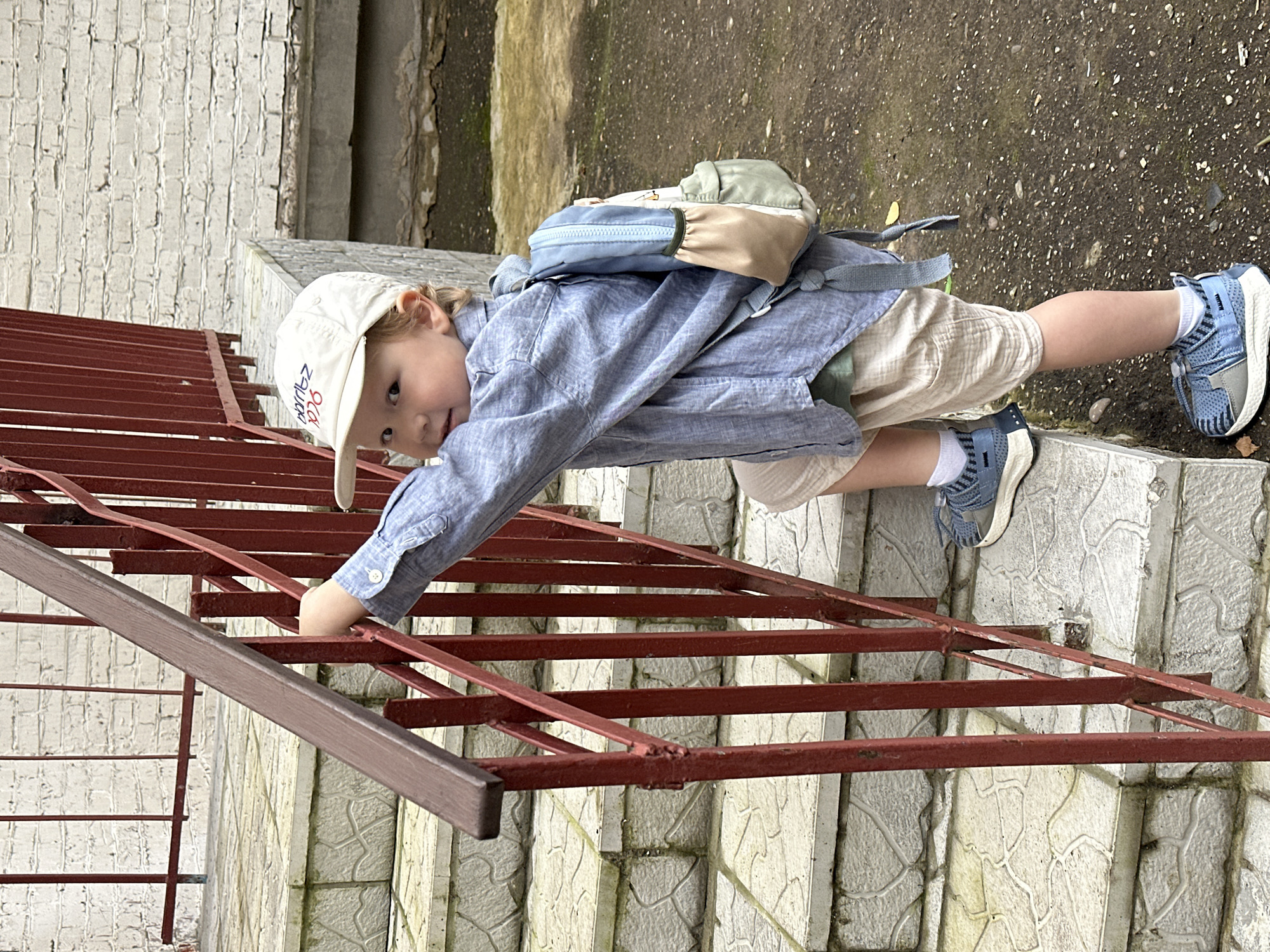 A young child wearing a light blue collared shirt, light beige shorts, a white baseball cap, and blue sneakers is climbing on a red metal railing alongside a concrete staircase. The child has a small, light-colored backpack with blue, tan, and green sections on their back. The child is looking towards the camera with a curious expression. The scene appears to be outdoors, with a textured wall behind the child and a concrete ground with scattered pebbles. The railing is angled, suggesting it's part of a staircase or balcony. The image is shot in a vertical orientation. The location context is Votkinsk, Russia. There is some visible text on the child's cap, which appears to be in Cyrillic script and possibly a logo or brand name.