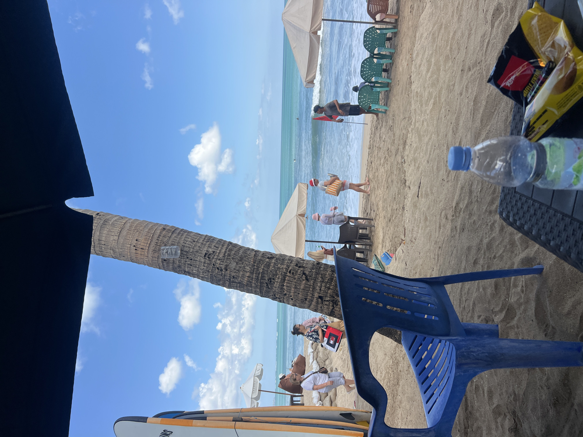 A group of people are on a sandy beach in Kuta, Indonesia. The scene is framed by a large dark umbrella in the foreground. In the foreground, a blue plastic chair is visible on the sand, and a plastic water bottle and a bag of chips are on a table. Behind the chair, a large palm tree stands tall. Several surfboards are leaning against the tree.
Further back, people are walking along the beach and near the water. Some are under umbrellas, and beach chairs are set up on the sand. The ocean is calm with small waves, and there are a few people in the water. A red flag is planted in the sand near a group of green chairs. The sky is blue with scattered white clouds.