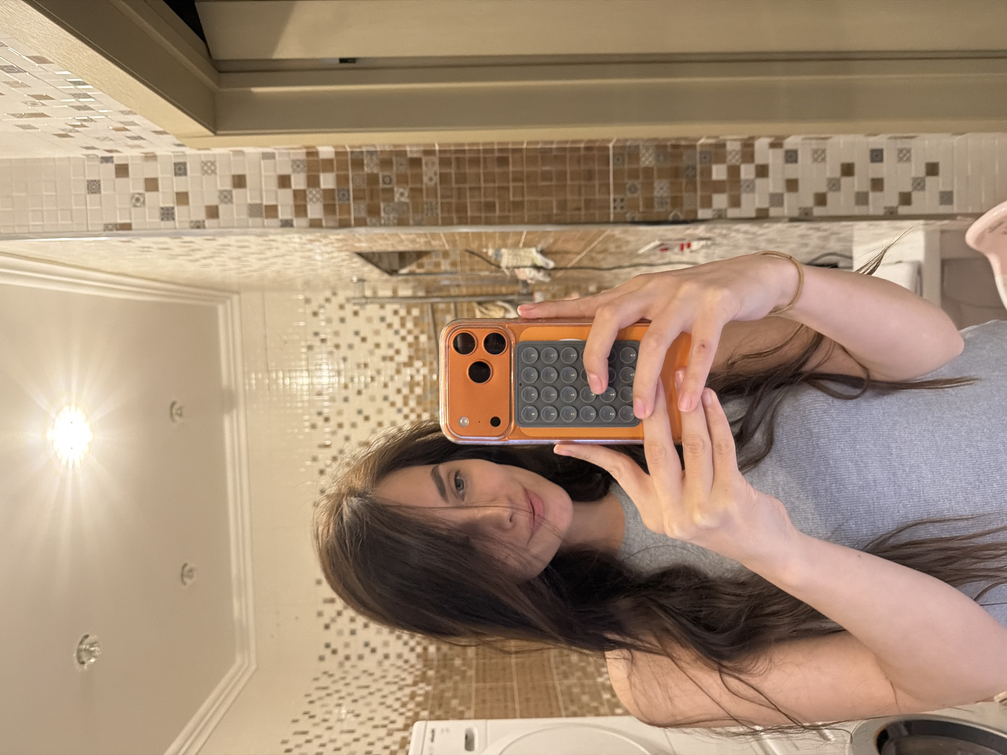 A young woman with long, dark, wavy hair is taking a selfie in a bathroom. She is wearing a grey tank top and has a delicate bracelet on her right wrist. Her phone, encased in an orange cover with a black circular button keypad, is held in her hands as she captures the reflection. The bathroom walls are tiled with a mosaic of cream, brown, and white squares. A washing machine is partially visible to her left. The ceiling features a prominent bright light fixture. The overall setting suggests a domestic environment.