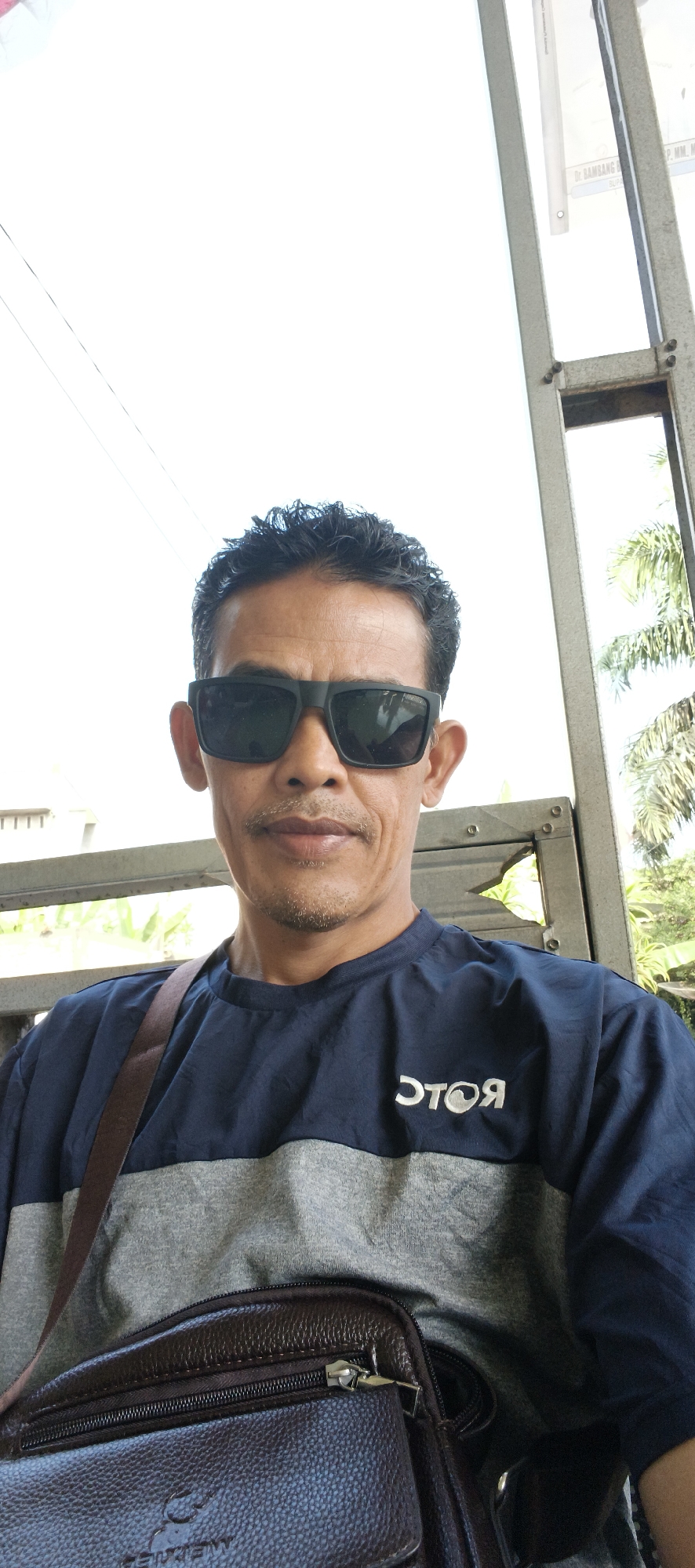 A man with dark, curly hair and sunglasses is seated outdoors, his upper body filling the frame. He is wearing a navy blue and gray t-shirt with a white logo on the right side. A brown crossbody bag is visible resting on his lap, with part of its strap draped over his shoulder. The man appears to be in a covered outdoor area, possibly a bus stop or waiting area, with a metal structure to his right and what looks like a fence or gate behind him. Lush green foliage and palm trees are visible in the background, suggesting a tropical or subtropical environment. The sky is bright and overcast. The overall scene is a casual, everyday moment captured in what appears to be a public space in Jambi Kecil, Indonesia.