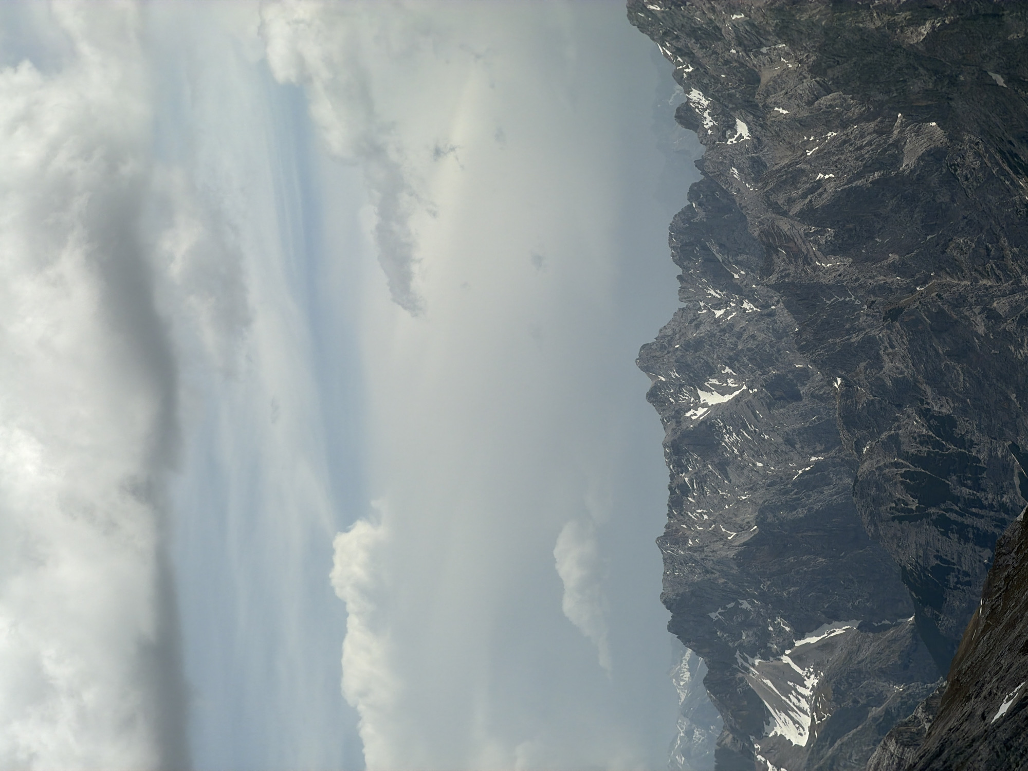 The image shows a landscape in Garmisch-Partenkirchen, Germany. On the right side, there is a rugged mountain range with visible rock formations and patches of snow. The left side of the image is filled with a cloudy sky that includes both dense gray clouds and areas of lighter, brighter sky. The orientation of the image is such that the sky appears on the left and the mountains on the right.