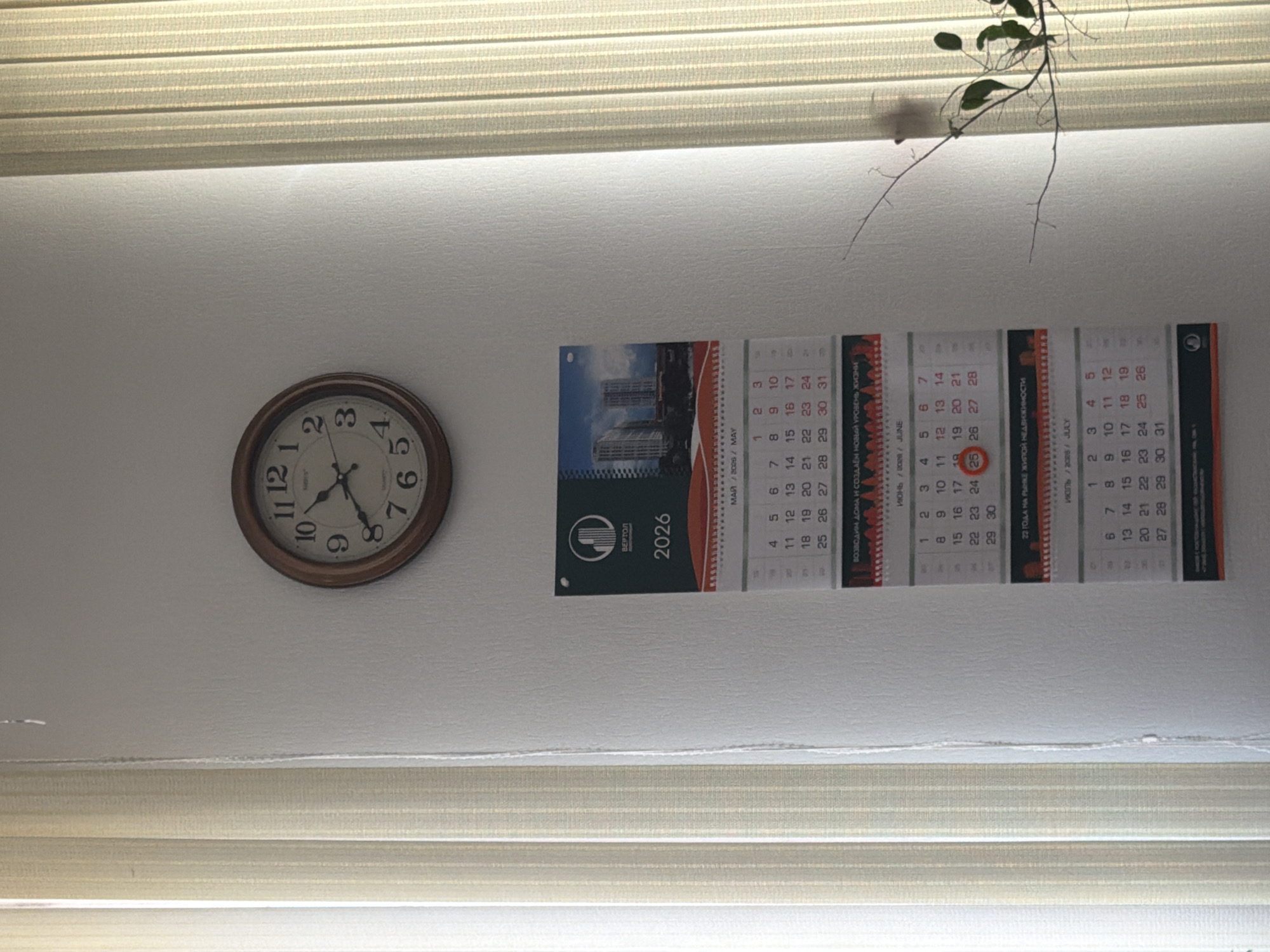 The image shows a wall with a clock and a calendar hanging on it. The clock is round with a wooden frame and Roman numerals. The calendar displays the months of May, June, and July of 2026. The top section of the calendar features an image of modern buildings and the logo of "ВЕРТОЛ" (Vertol). Below this, the calendar shows the days and dates for each month. The image is well-lit, suggesting an indoor setting with natural light coming from vertical blinds on the left side of the frame. A thin branch with a few green leaves extends into the frame from the right, partially obscuring the calendar. The wall appears to be painted a light color, and the overall scene is tidy and well-maintained.