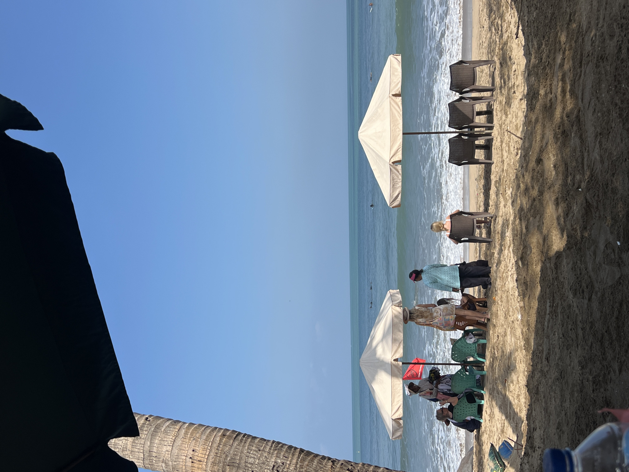 In Kuta, Indonesia, a scene unfolds on a sandy beach with the ocean in the background under a clear blue sky. A large palm tree trunk is visible on the left side of the frame, partially obscured by a dark umbrella. In the mid-ground, two large, light-colored beach umbrellas provide shade for a group of people and several plastic chairs.
Under the first umbrella, a vendor dressed in traditional clothing appears to be interacting with two women seated on chairs. One woman is wearing a hat, and the other is partially visible. A red flag is planted near the vendor. Next to them, three green plastic chairs are empty.
Under the second umbrella, a woman in a light-colored dress stands facing away from the camera. To her right, a person is seated in a dark chair, also facing away. Further to the right, three dark plastic chairs are arranged in a row, two of which are occupied by individuals facing the ocean. In the distance, several surfers can be seen in the turquoise water, which has gentle waves breaking on the shore. The foreground of the image is sandy, with some shadows cast by the umbrellas and palm tree. A blue plastic bottle cap is visible in the lower left corner of the frame.