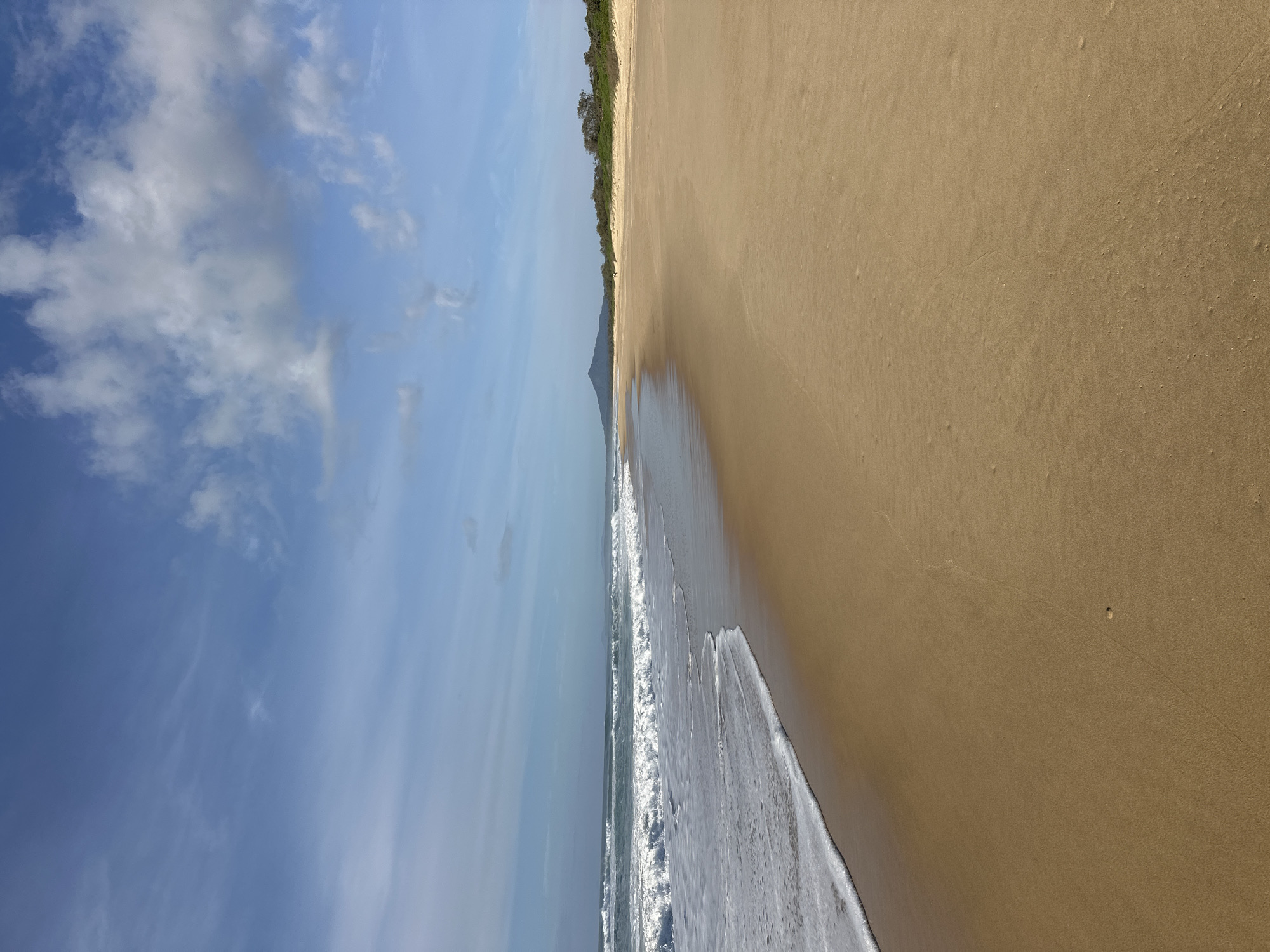 The image depicts a wide, sandy beach in Mylestom, Australia, under a partly cloudy blue sky. The ocean waves are breaking gently on the shore, creating a foamy white edge. The sand is a golden-brown color, with some slight ripples and small indentations visible. In the distance, along the coastline, there are trees and a conical mountain. Two figures can be seen walking on the beach in the middle distance, appearing as small silhouettes against the sand and vegetation. The overall scene is tranquil and natural, showcasing a typical Australian coastal landscape.