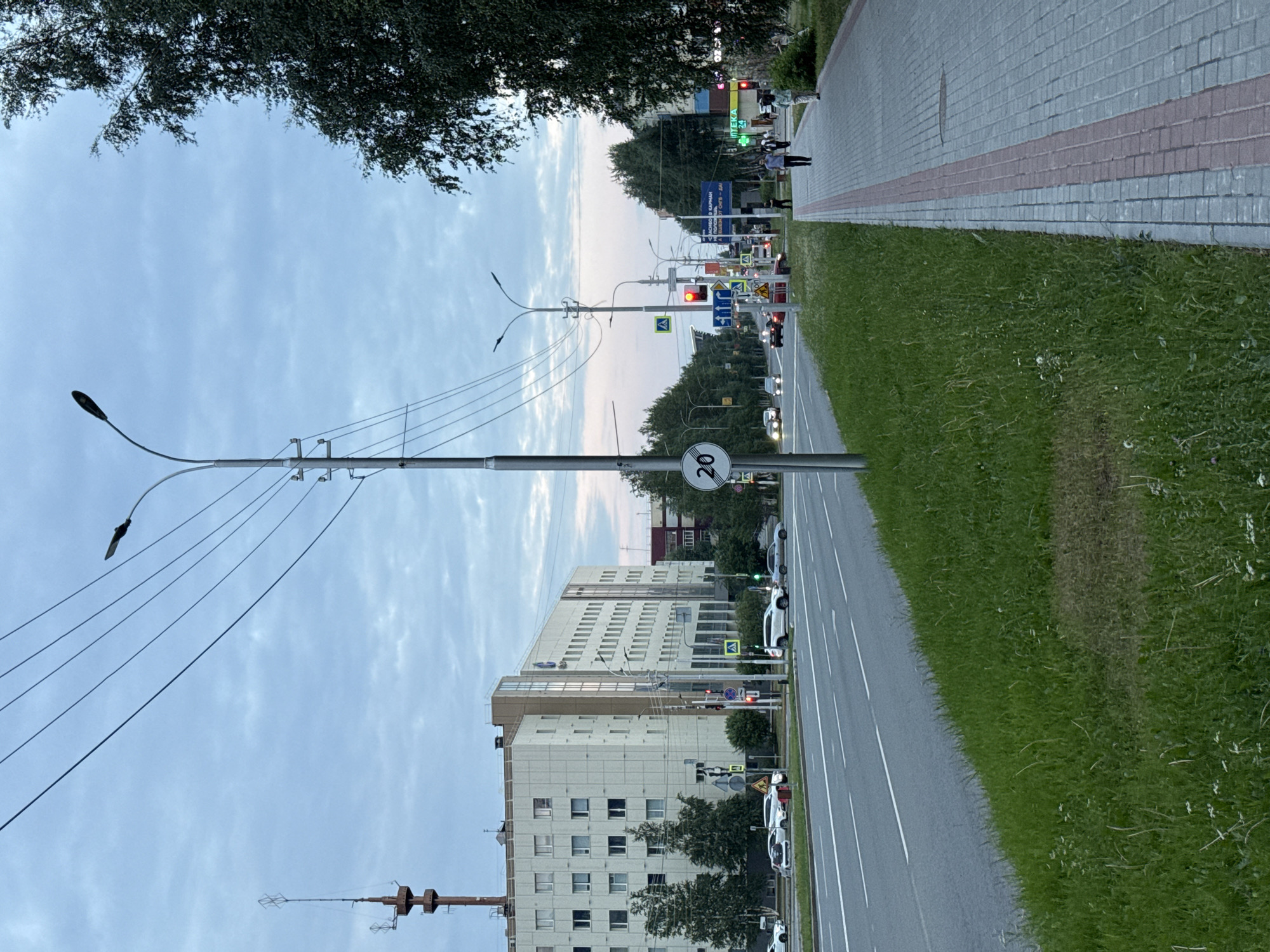 The image captures a street scene in Surgut, Russia, likely during the late afternoon or early evening, given the soft, diffused lighting and the overcast sky. The perspective is from the side of a road, looking down a sidewalk and then across the street.
On the right side of the image, a well-maintained sidewalk made of light-colored pavers with a contrasting red stripe runs parallel to the road. Adjacent to the sidewalk is a strip of vibrant green grass, which appears neatly mown. The road itself is paved and marked with white lane lines. Several cars are visible on the road, traveling in the same direction as the viewer's perspective. A speed limit sign displaying "20" is prominently featured on a pole by the sidewalk.
On the left side of the street, several multi-story buildings dominate the background. These appear to be modern office or residential buildings, with numerous windows. A tall lamppost with several wires extends from a pole on the left side of the image, its light fixture extending over the sidewalk. Further down the street, traffic lights and various road signs are visible, indicating a busy intersection. There are also trees lining the street in the distance.
A few people are visible on the sidewalk in the distance, walking. One person in the foreground on the sidewalk is partially obscured by a tree. The overall atmosphere is calm and ordinary, depicting everyday urban life in a Russian city. There is no clear indication of any specific event or strong emotion.