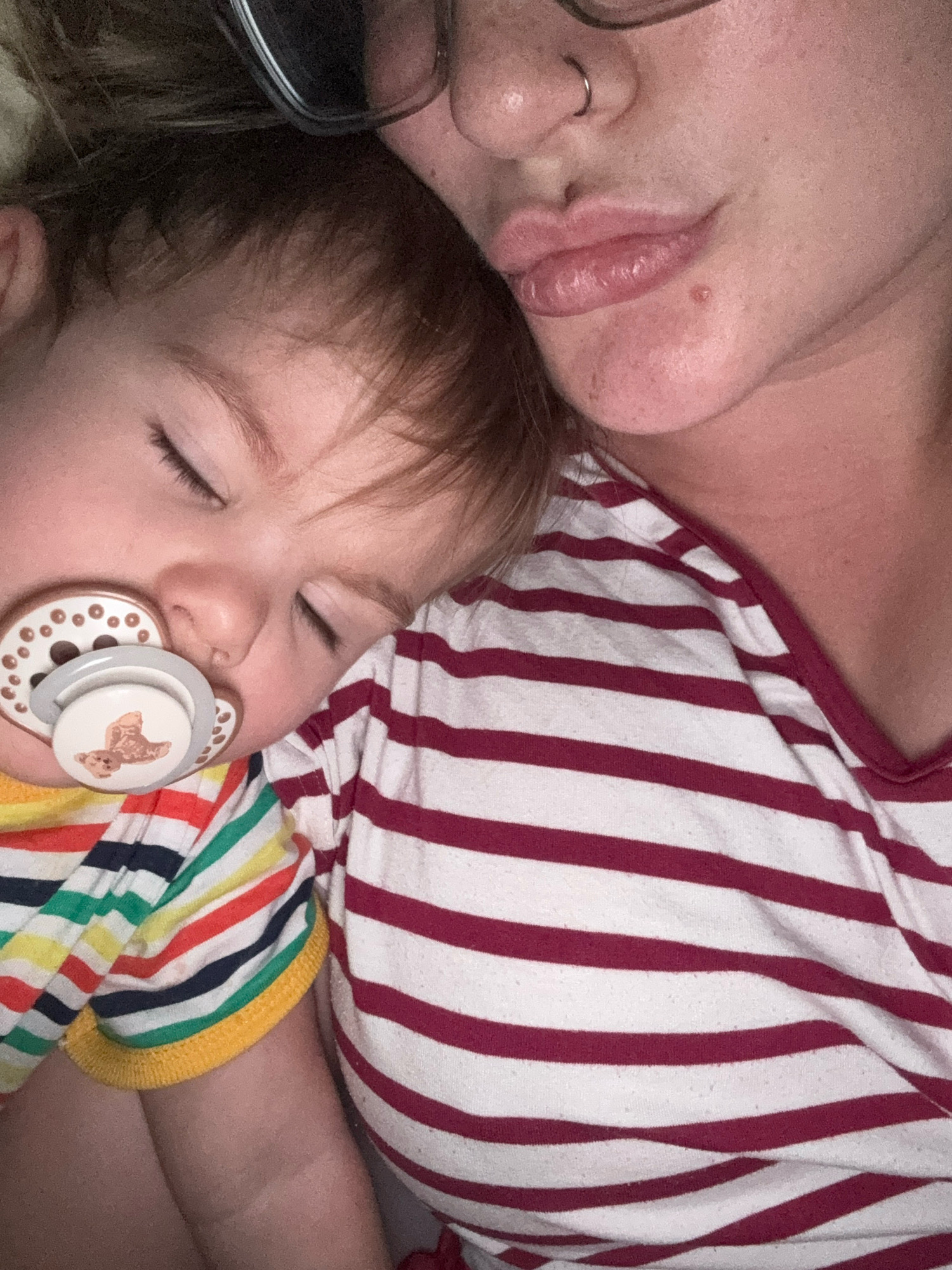 The media file shows a close-up of a baby sleeping on a person's shoulder. The baby, with eyes closed and a pacifier in its mouth, is wearing a brightly colored striped shirt with yellow trim. The person's shoulder and upper chest are visible, clad in a red and white striped shirt. The person is wearing glasses and has a nose ring. The lighting suggests it is nighttime or indoors. The scene evokes a sense of comfort and intimacy. No text is visible in the image. The location context is Stavropol, Russia.