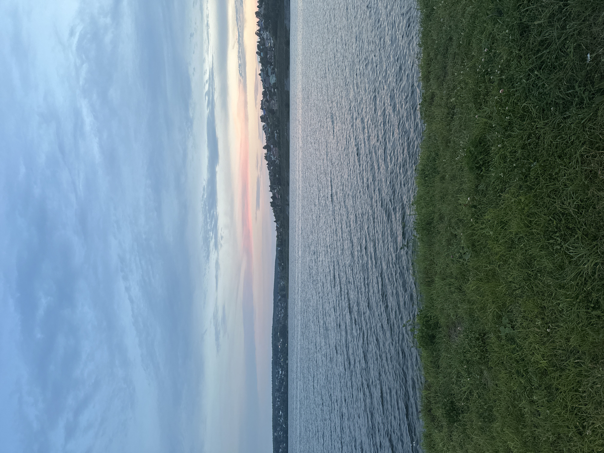 The image depicts a scenic view of the Lysva River in Lysva, Russia, likely during either sunrise or sunset. The sky is a soft gradient of blue and pink hues, with scattered clouds. The river occupies the middle ground, its surface rippled by a gentle breeze. Along the far bank, a picturesque settlement with houses and trees is visible, nestled on a gentle slope. The foreground is dominated by a lush green grassy embankment. The overall impression is one of tranquility and natural beauty.