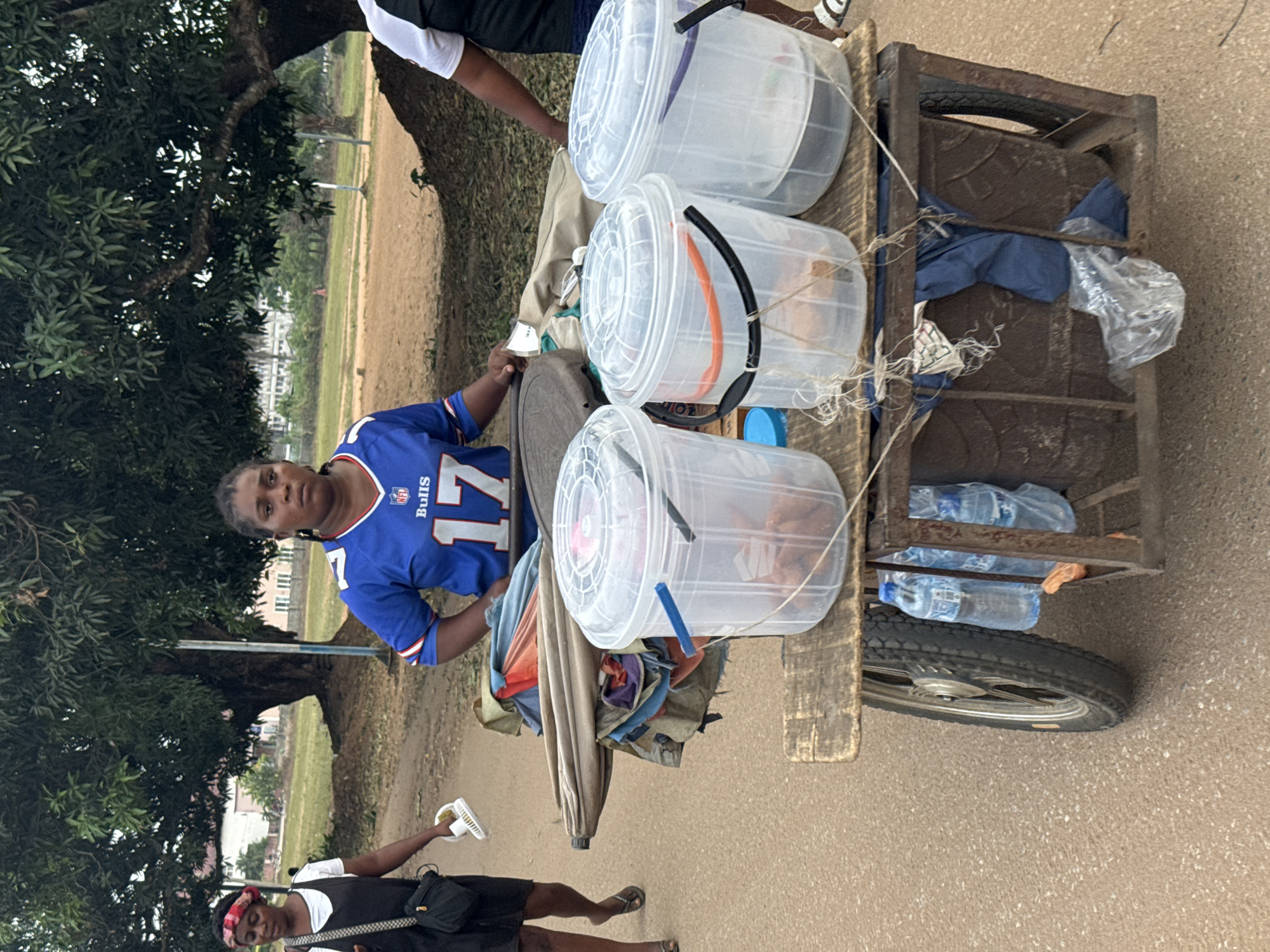 The scene depicts an outdoor setting in Ihiagwa, Nigeria, featuring three individuals and a mobile vendor's cart on a light brown, unpaved surface. In the central foreground, a woman wears a blue NFL "Bills 17" jersey and stands behind a two-wheeled wooden and metal pushcart. She has braided hair and a neutral expression, with her hands resting on the cart's surface.
The cart's wooden top supports three large, clear plastic buckets with clear lids, each containing brown-colored solid items, likely food. The buckets have handles: one blue, one orange, and one black/purple. Behind these buckets, several folded items resembling umbrellas or tarps, and assorted fabrics, are stacked. Underneath the wooden platform, within the cart's metal frame, several plastic water bottles, blue fabric, and a clear plastic bag are visible.
To the left, another woman walks away from the camera. She wears a black sleeveless top over a white shirt, a dark skirt or shorts, and dark flip-flops. Her head is adorned with a red and white patterned headband. She carries a small white portable fan in her right hand and a black shoulder bag across her body. On the far right, a third person is partially visible, showing their torso in a black and white top and light-colored sandals.
The background consists of dense green foliage from large trees, open grassy areas, and distant multi-story white buildings. The ground appears to be dirt or an unpaved road. The lighting indicates daylight conditions.
