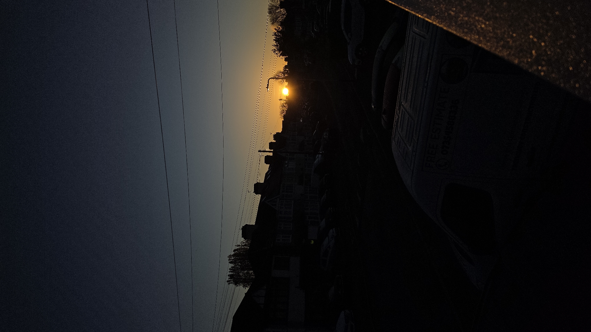 The image depicts a twilight or sunset scene in what appears to be a residential area. Silhouetted houses and trees line the street, with a bright light, possibly a street lamp or the setting sun, shining through the trees. Power lines run across the left side of the frame. A vehicle is parked on the right, with the text "FREE ESTIMATE" and a phone number partly visible on its side. The overall lighting is dim, obscuring details. There is no evidence to establish the location as London, United Kingdom.
