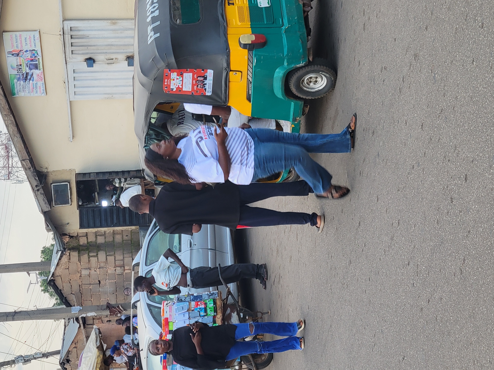This image captures a street scene in Barakin Lerawi, Nigeria, featuring several people and objects against a backdrop of buildings and a road. In the foreground, a woman in a white and blue striped shirt and blue jeans is seen from the chest down, holding a smartphone. She appears to be standing next to or leaning on an auto-rickshaw, which is a common mode of transportation in the region. The auto-rickshaw is primarily green and yellow with "DELUXE" printed on its side.
Behind the woman, two men are partially visible. One is wearing a black shirt and dark pants, and the other is in a white t-shirt and dark pants, seemingly interacting with a cart laden with various items, possibly for sale. Further in the background, more people are present near a white car and a structure that appears to be a shop or dwelling. The overall scene suggests a bustling marketplace or a busy street where daily activities are taking place.
A prominent piece of text visible on the auto-rickshaw reads "PT 282". On the side of a building to the left, a sign for "GENYA PRINTING" advertises services like "Program of Events," "Jupyter's Receipt book," "ProactiBook Binding," "Type Setting," "Calenders," "Fe and Jumas," and "DIGITAL PRINTING" as well as "General Contractive." The lighting suggests it is daytime, likely late afternoon given the warm tones and shadows. The people's expressions are not clearly discernible, but the casual nature of the interactions suggests a typical day.