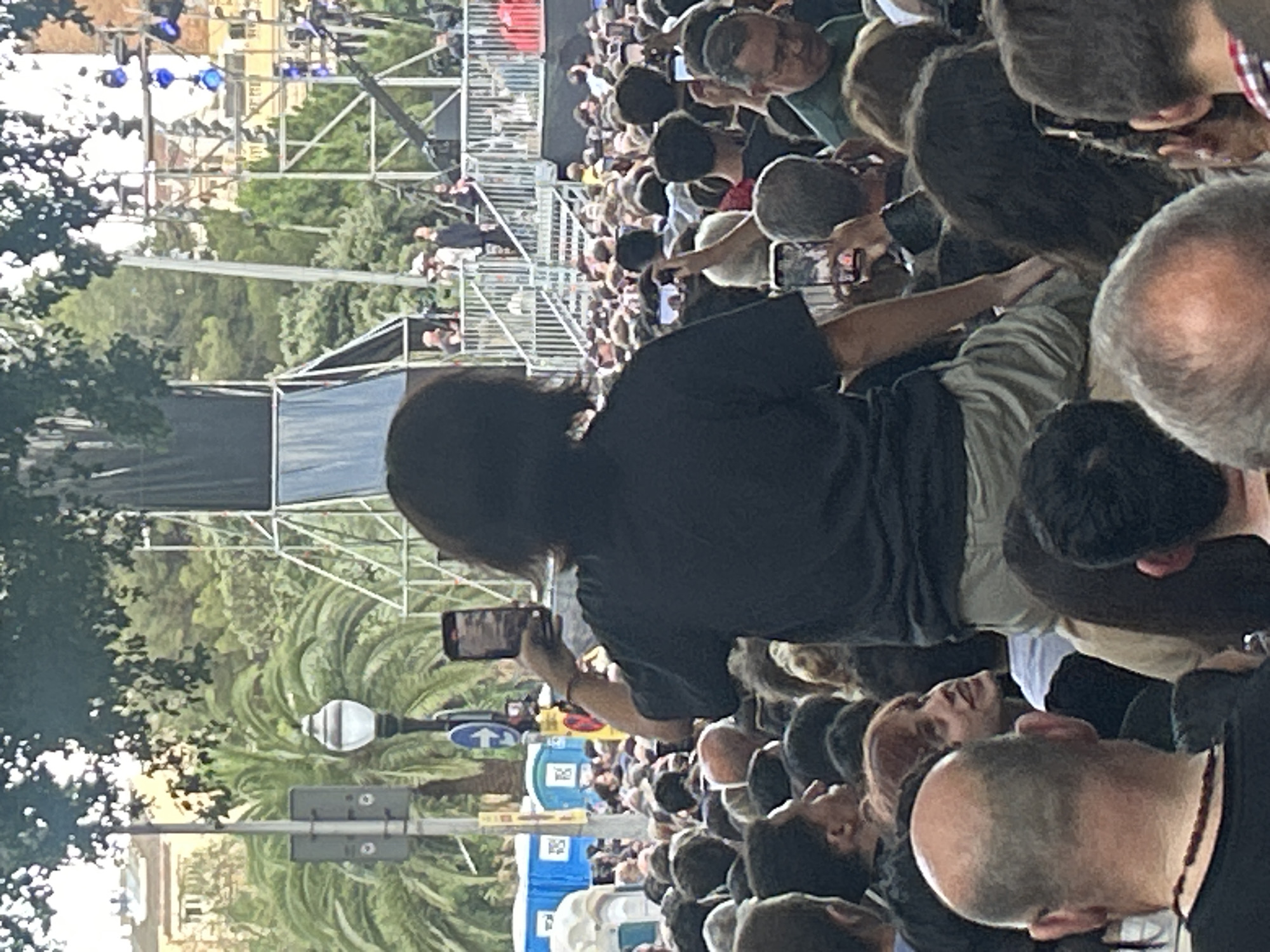 The image shows a crowd of people gathered outdoors, possibly for an event. In the foreground, a person is sitting on someone's shoulders, holding up a smartphone to take a picture or video. The person sitting on shoulders has dark hair and is wearing a dark t-shirt and light-colored shorts. The crowd is dense, with many people looking in the same direction, towards what appears to be a stage or performance area in the background. There are palm trees and buildings visible, suggesting an urban setting. The presence of portable toilets in the background also indicates a public event. The lighting suggests it is daytime. The architectural style of the buildings and the presence of palm trees could indicate a Mediterranean climate. Given the context of large outdoor gatherings, it is plausible this scene is in Barcelona, Spain, a city known for its vibrant culture and public events.