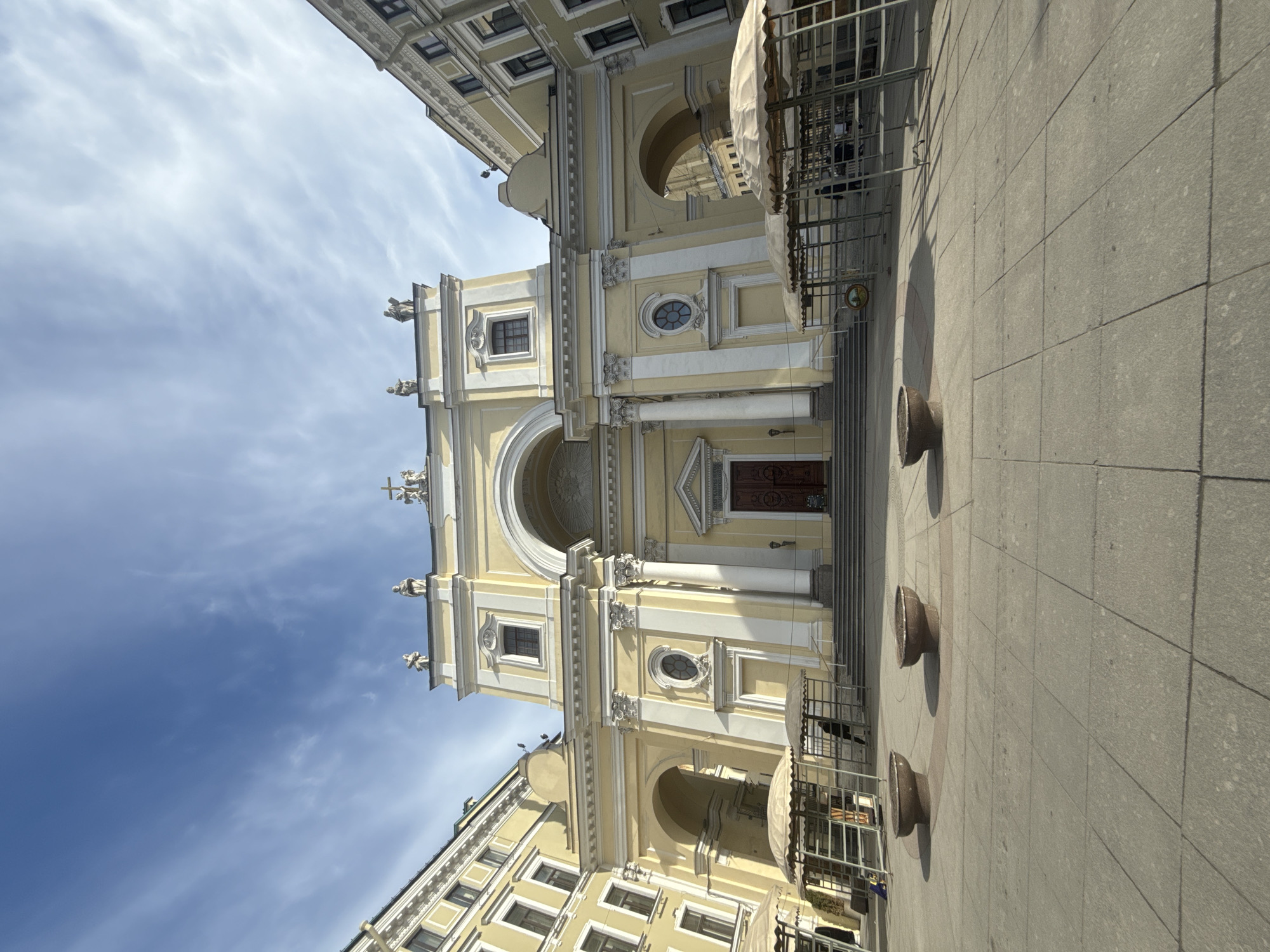 The image depicts a grand, ornate church or cathedral, rendered in a pale yellow color with extensive white architectural details, under a bright blue sky with wispy white clouds. The building features a prominent central arched entrance with dark wooden double doors, flanked by large white columns and pilasters. Above the main entrance is a large archway with intricate decorative molding. The facade is adorned with multiple rectangular and circular windows, all framed in white, and features several statues positioned along cornices and pediments, including a cross at the apex of the left pediment. The overall architectural style appears to be Baroque or Neoclassical, characteristic of structures found in Saint Petersburg, Russia.
To the right side of the main building, scaffolding and white tarps are visible, suggesting ongoing renovation or construction work. The foreground consists of a spacious paved plaza or sidewalk made of large, light grey rectangular tiles. Three low, dark-colored, round stone planters or bollards are arranged on the plaza, casting distinct shadows to their right, indicating a strong light source, likely the sun, from the left. Other multi-story buildings, consistent in style and color, are partially visible on the left and right edges of the frame, suggesting an urban environment. The scene is devoid of people or discernible activity, presenting a quiet, sunny daytime view. No visible text is present in the image.