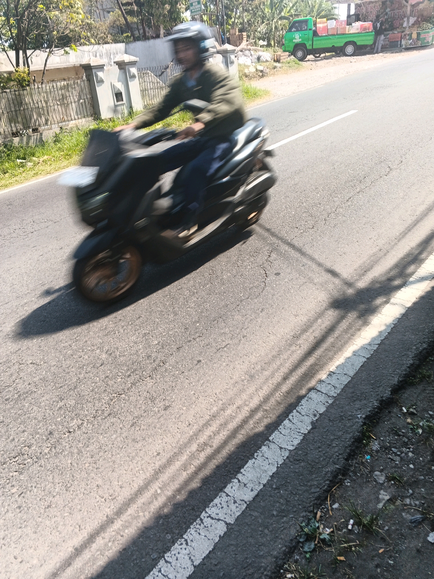 In Kenteng, Indonesia, a man wearing a helmet is riding a dark-colored scooter down a paved road. The scooter is in motion, as indicated by the blur. The man is wearing a green jacket and dark pants. Beside the road, a light green pickup truck is parked, loaded with boxes. In the background, there is a low wall and some vegetation. The sun is casting shadows across the road.