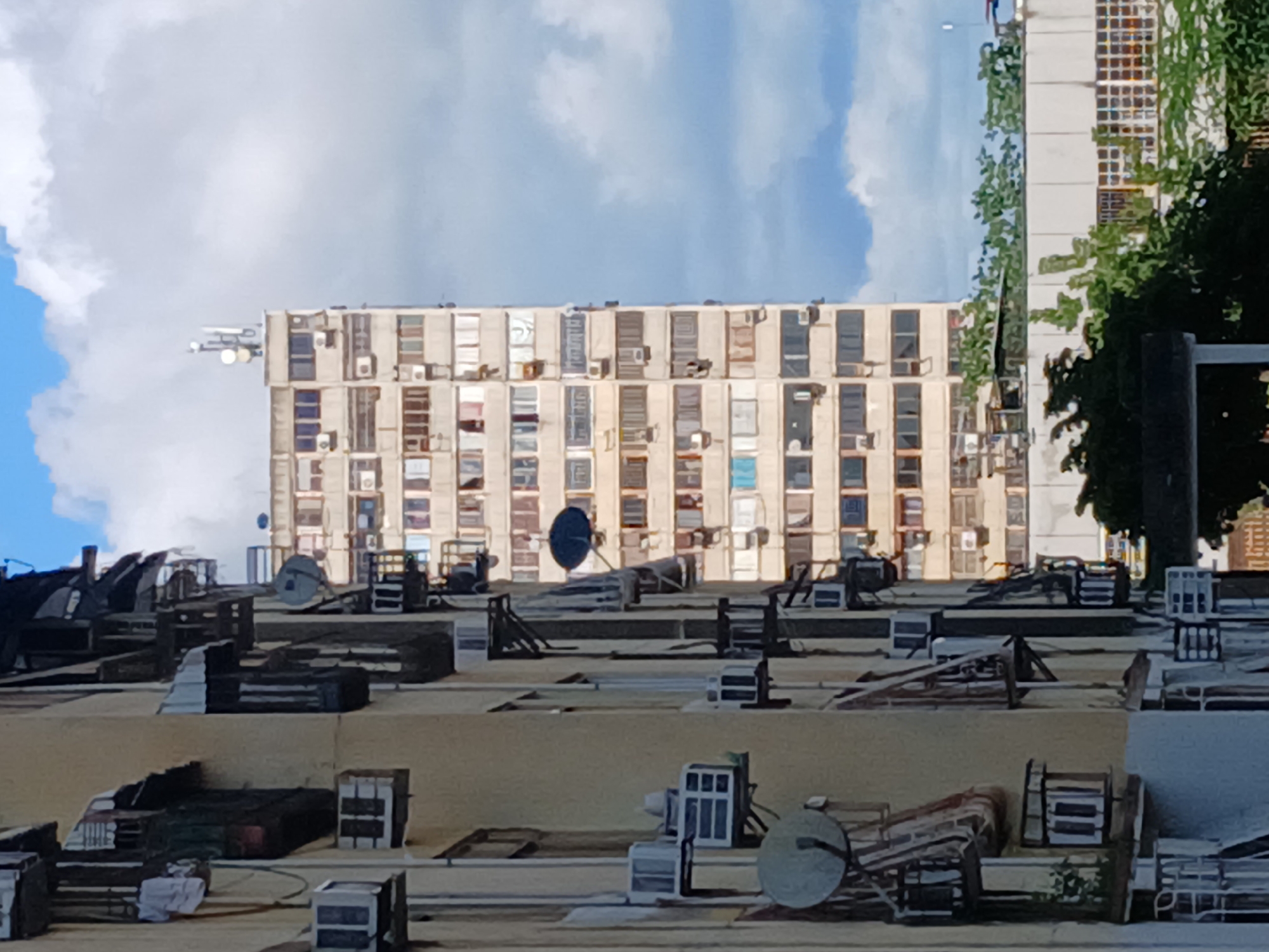 The image depicts a view of apartment buildings in Puerto Ordaz, Venezuela. In the foreground, a pale yellow apartment building with visible air conditioning units mounted on its exterior is partially obscured. Directly behind it, a taller, light-colored concrete high-rise apartment building is prominently featured. This building has numerous windows and appears to be densely populated with air conditioning units and satellite dishes. The sky above is partly cloudy with blue patches, indicating daytime. The scene is urban, characterized by dense residential architecture. No people are clearly visible in the image.